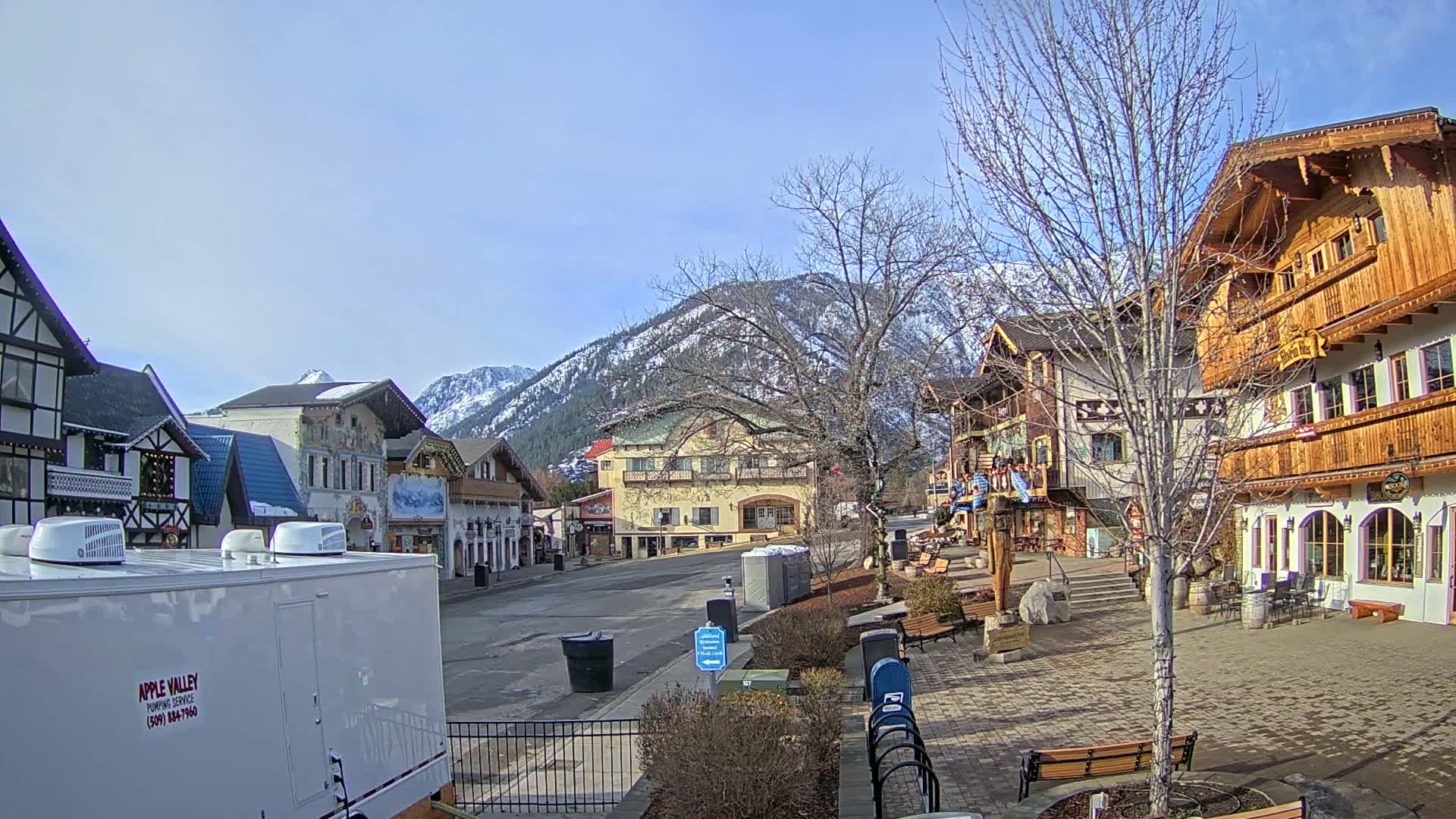 A sunny day in a village with European-style architecture, nestled in a valley with snow-capped mountains in the background.