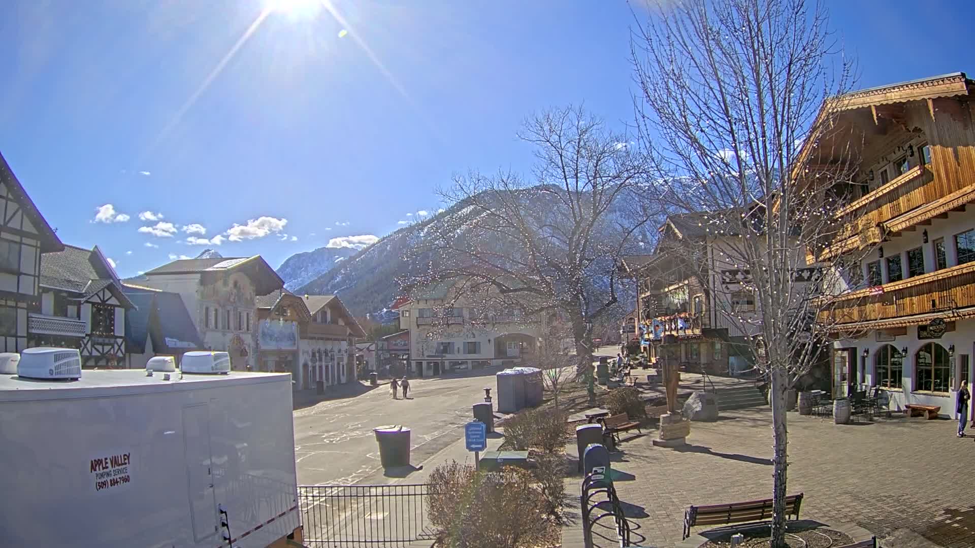 A sunny day reveals a village square with several buildings in a European style, situated at the foot of a snow-capped mountain range.