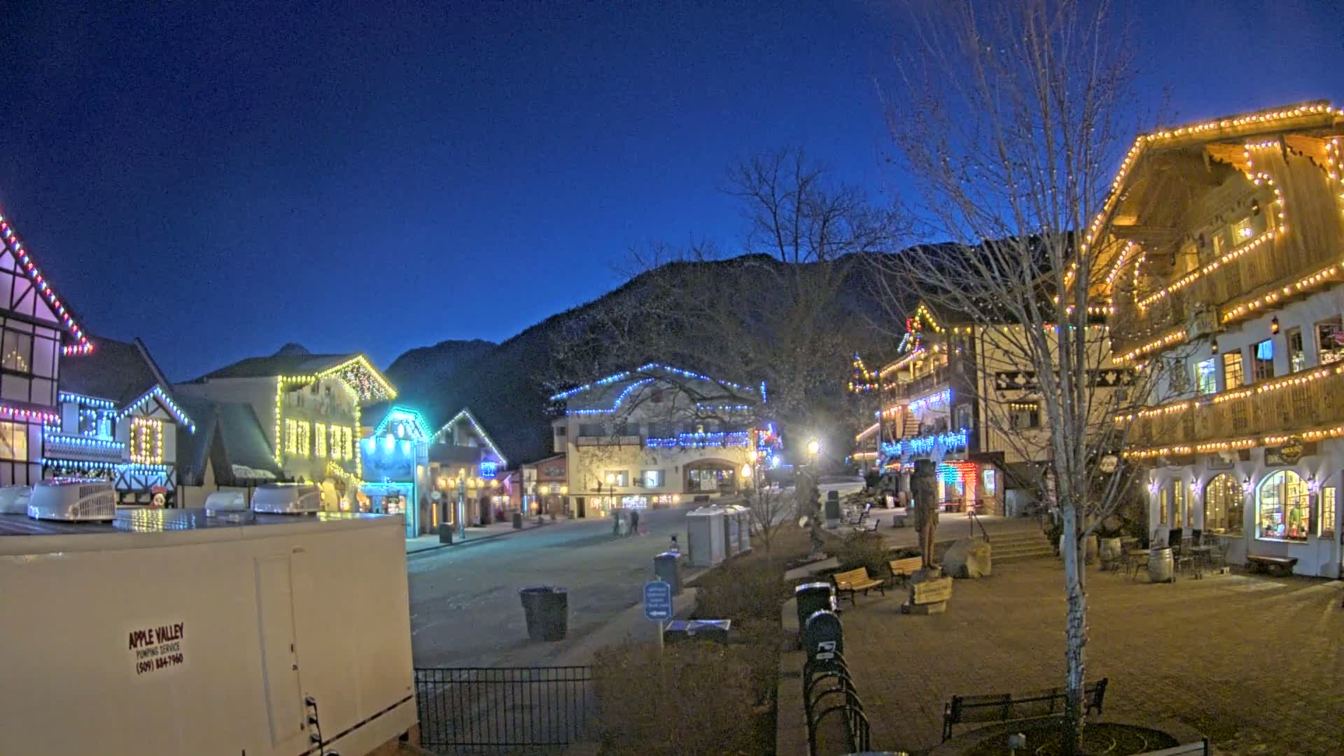 A nighttime scene of a village square with buildings decorated in colorful string lights, under a clear dark blue sky.