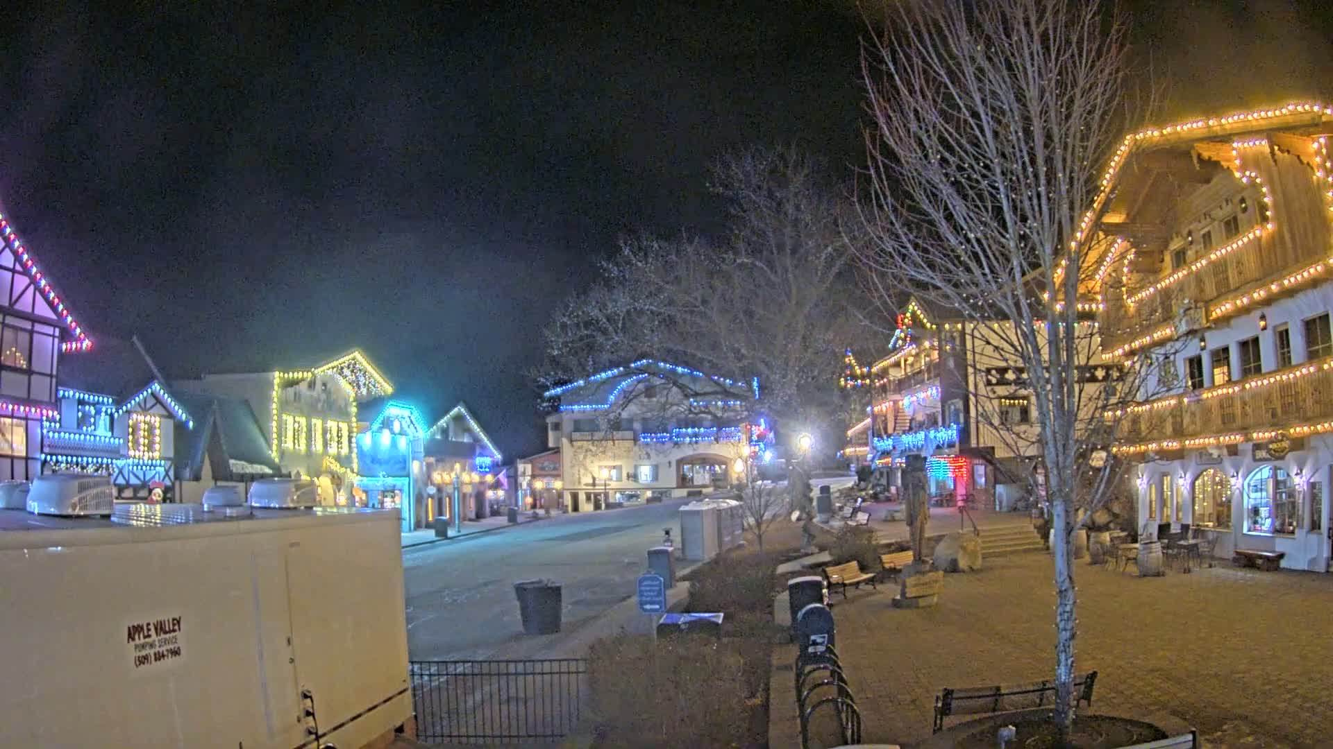 A nighttime scene of a town square adorned with Christmas lights, featuring several buildings with festive decorations and sparsely populated, snow-covered ground.
