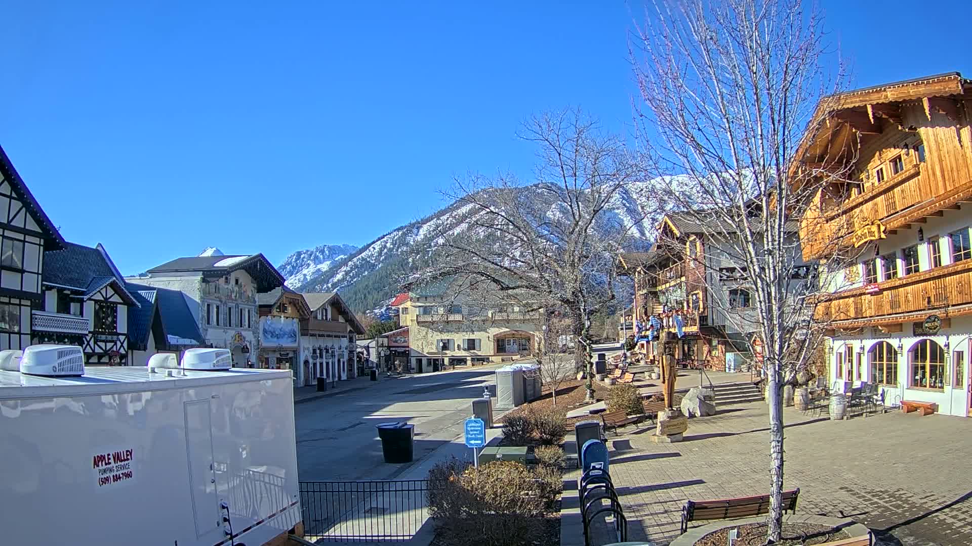 A snow-capped mountain range is visible in the background of a town square with several buildings in various architectural styles, under a clear blue sky.