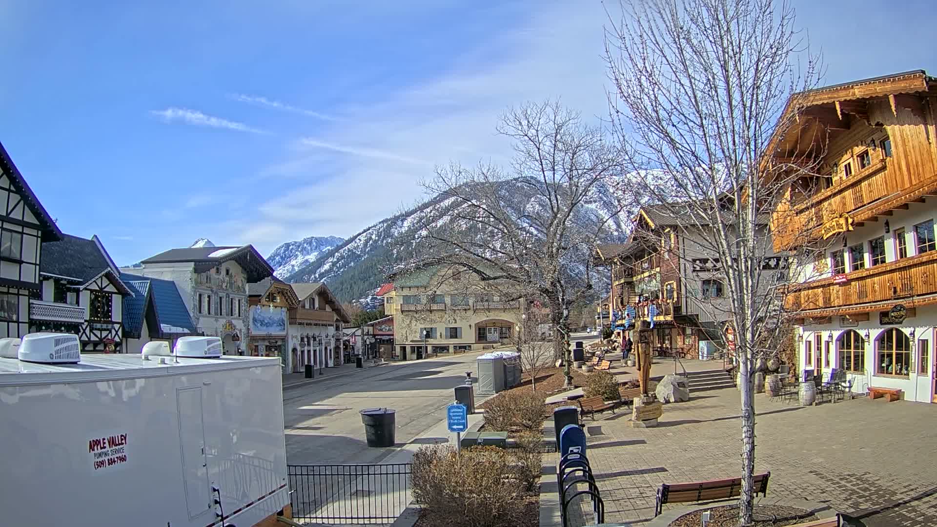 A quaint town square with buildings in a Bavarian style sits under a clear blue sky, with snow-capped mountains in the background.