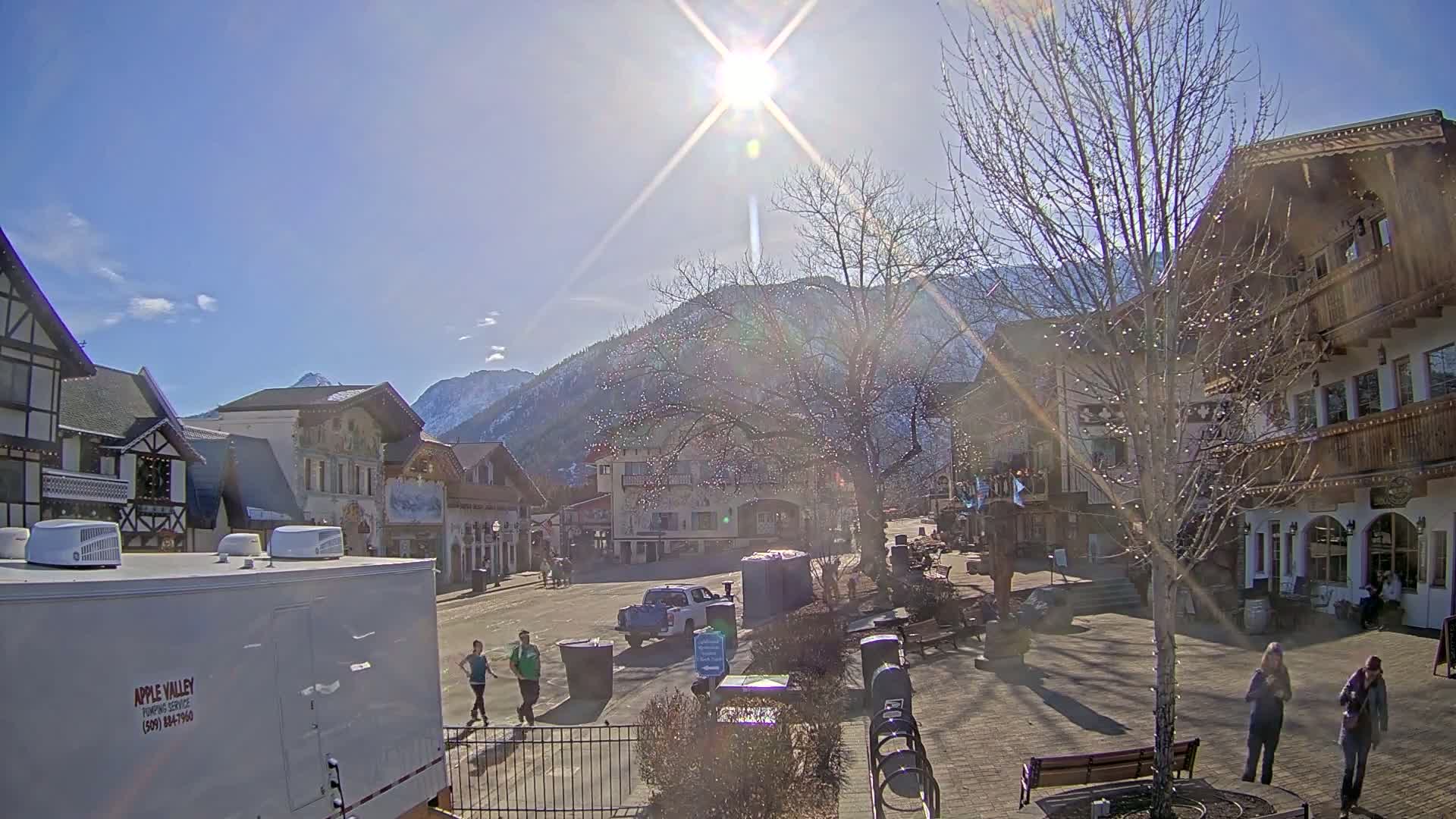A sunny day in a European-style village square, with several people walking and a mountain range in the background.