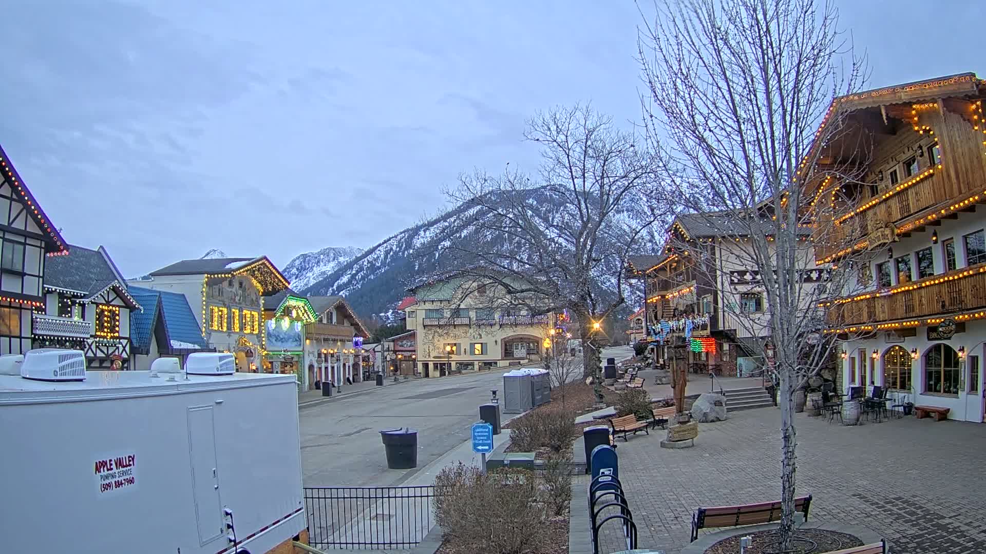 A mostly empty town square with quaint, festively lit buildings, under a partly cloudy sky, is nestled at the base of snow-dusted mountains.