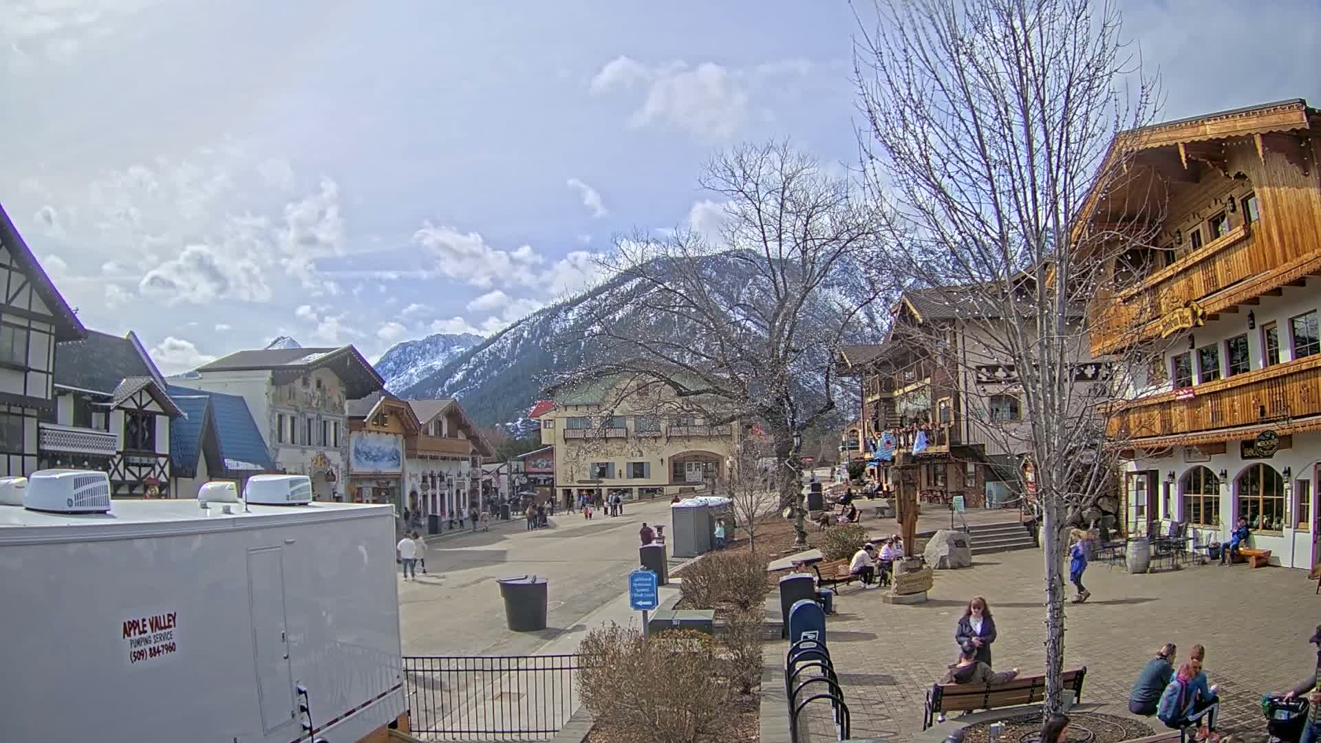A partly cloudy day reveals a Bavarian-style village square surrounded by shops and buildings, nestled at the base of snow-capped mountains.