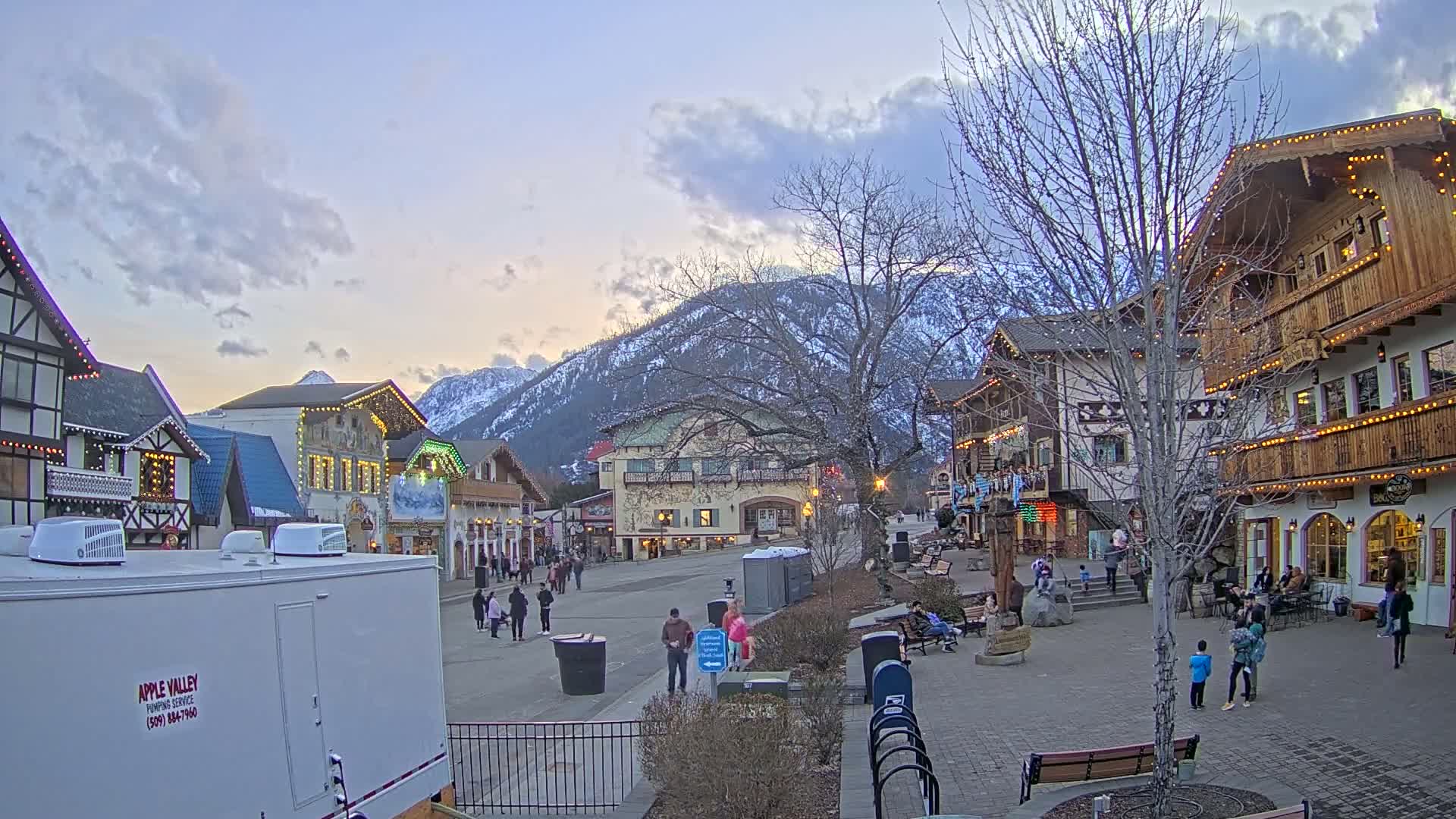 A town square with Bavarian-style buildings is populated with a few pedestrians under a partly cloudy twilight sky, with snow-capped mountains in the background.