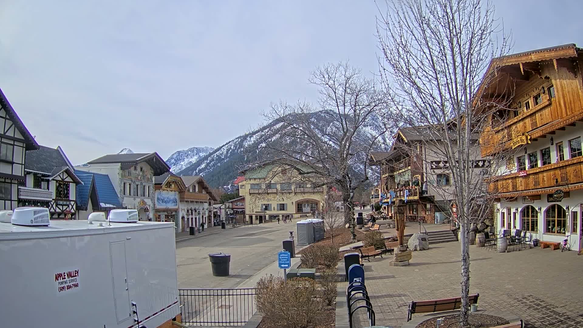 A town square with several Bavarian-style buildings sits in front of snow-capped mountains under a mostly cloudy sky.