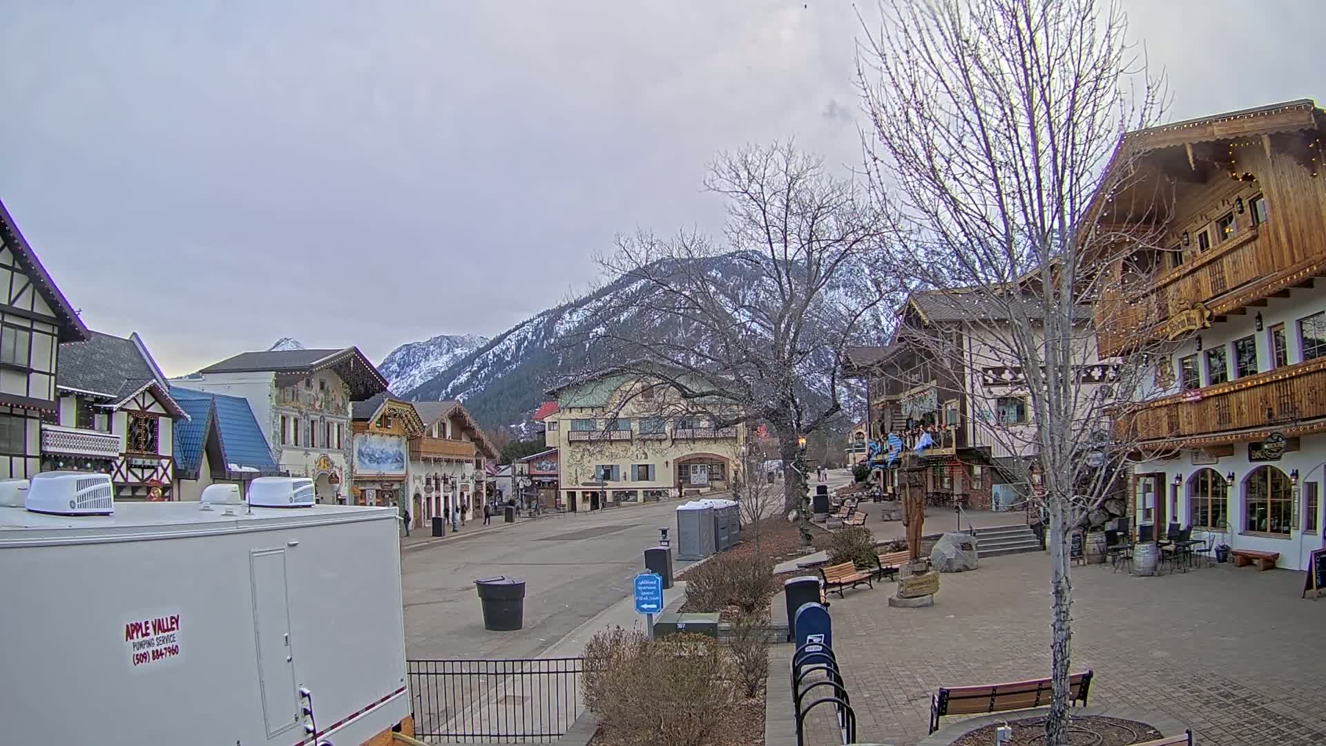 A mostly empty village street with alpine-style buildings and a mountain backdrop under an overcast sky.