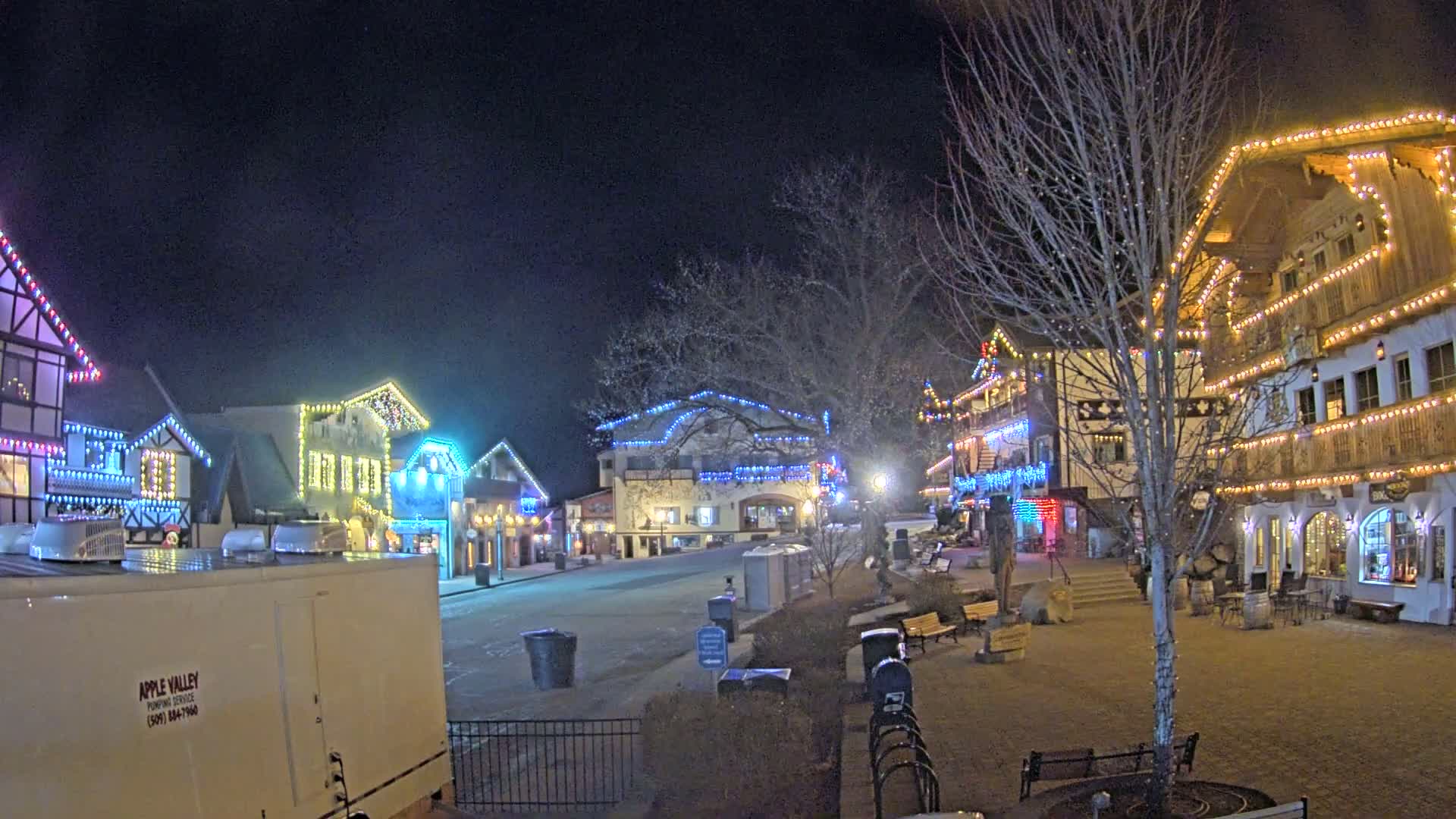 A town square at night, illuminated by strings of colorful lights on buildings, with bare trees and a few benches scattered throughout.