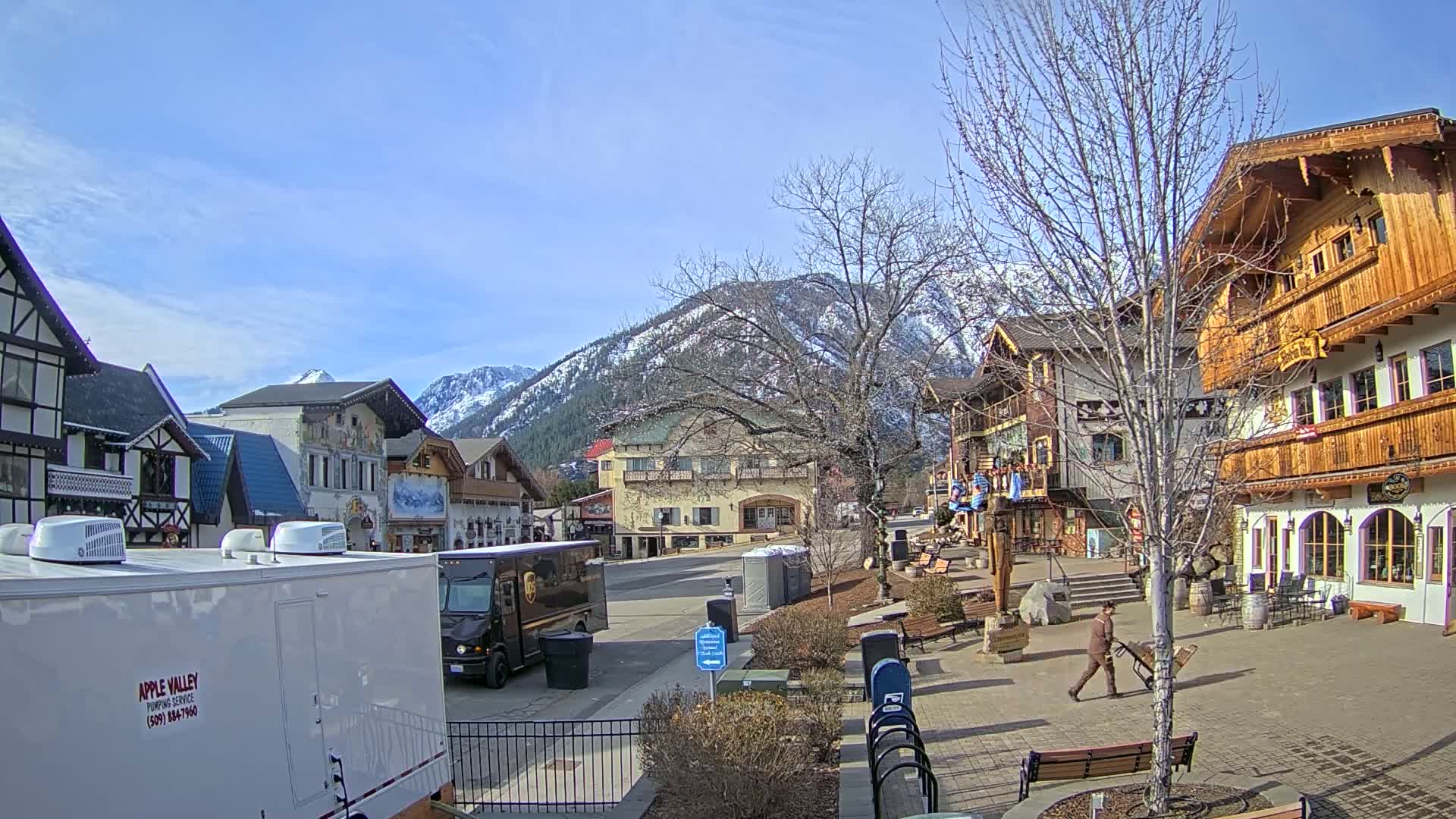 A sunny day reveals a quaint village square nestled in a valley surrounded by snow-capped mountains, with several buildings exhibiting European-style architecture.