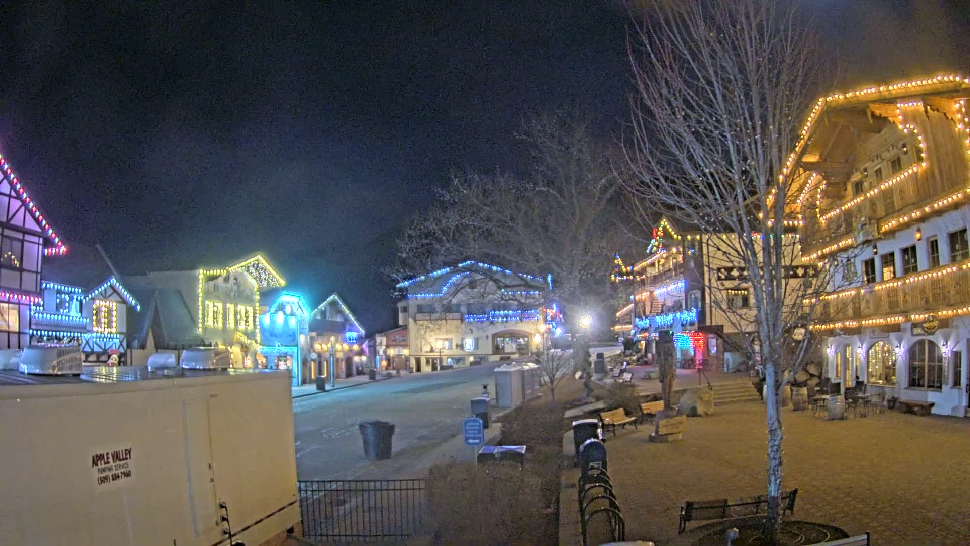 A nighttime scene of a town square with several buildings decorated with colorful Christmas lights, under a clear, dark sky.