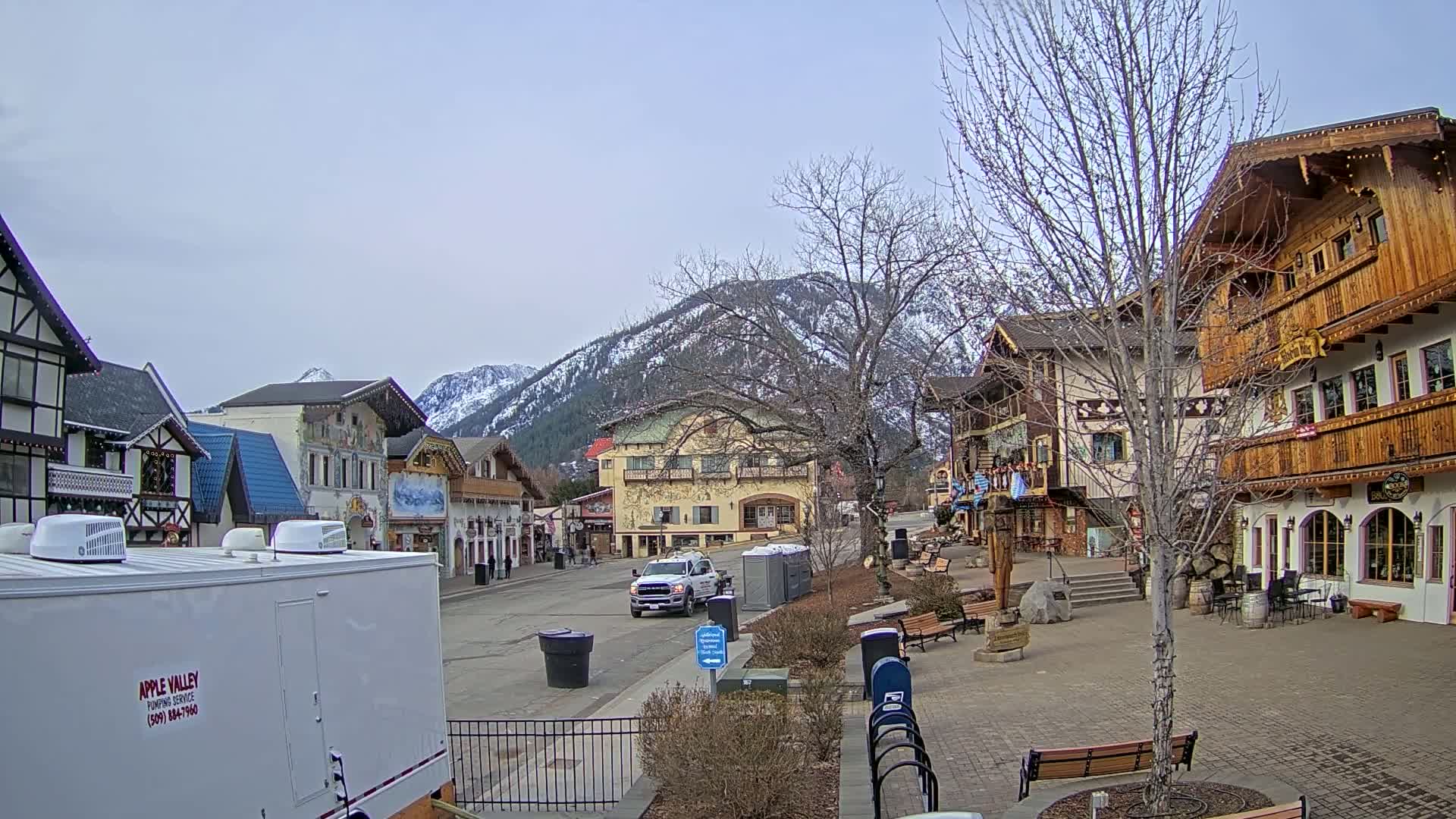 A village square with several buildings in an alpine style, under an overcast sky, is nestled against a snow-capped mountain range.