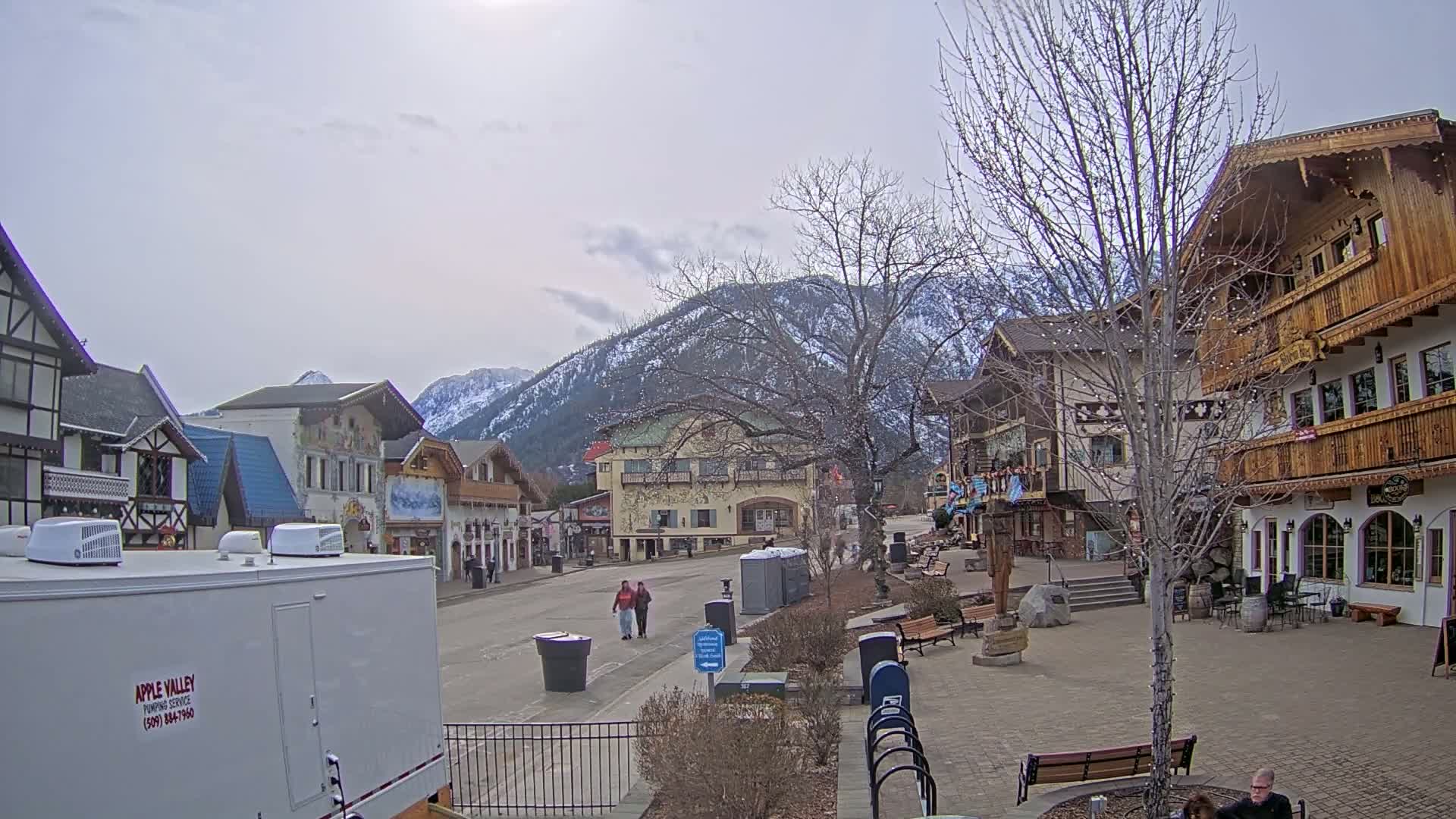 A mostly empty town square with snow-dusted mountains in the background under a cloudy sky features several Bavarian-style buildings and a few pedestrians.