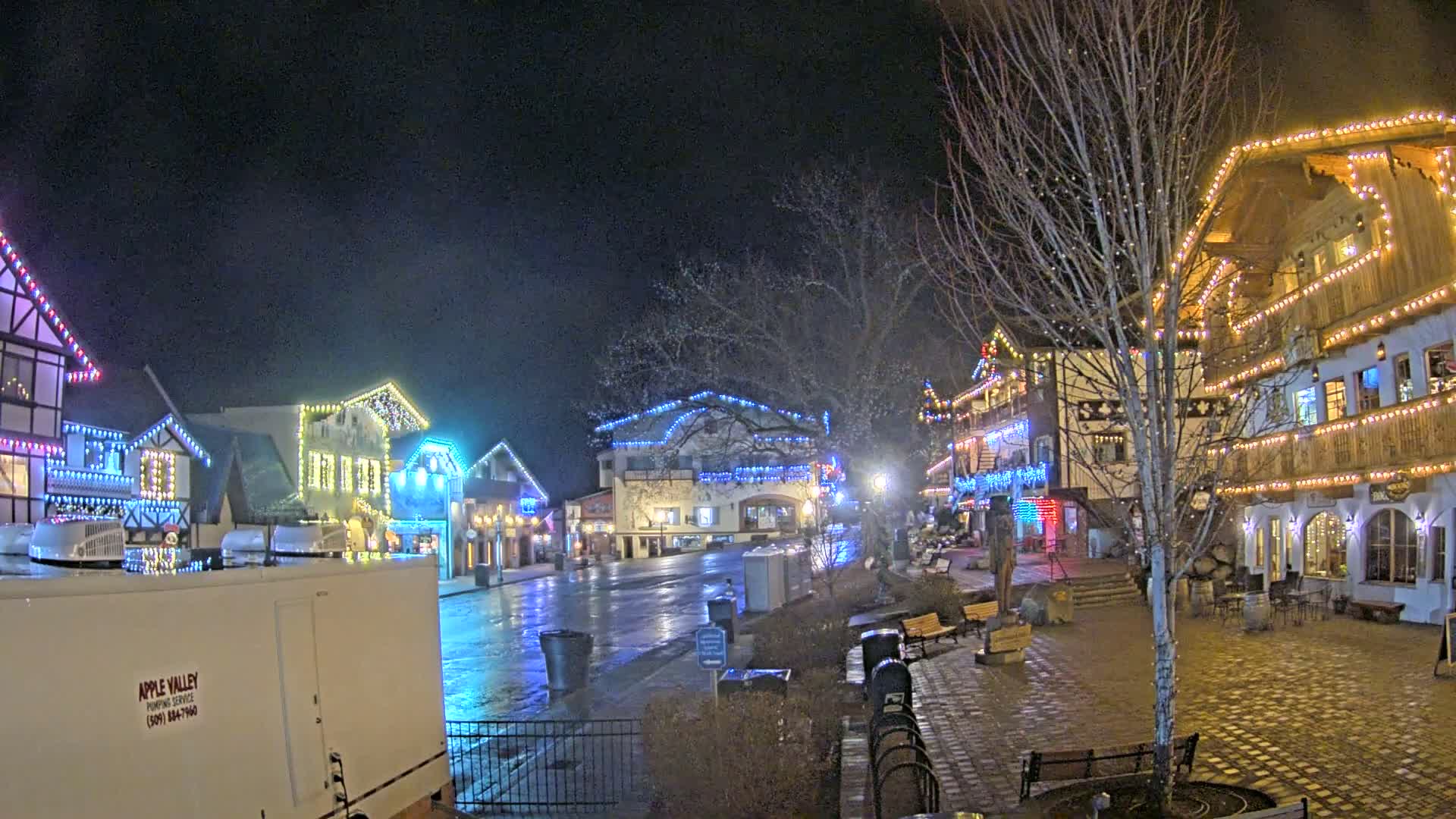 A town square at night, decorated with colorful Christmas lights, is wet from a recent rain or snow.
