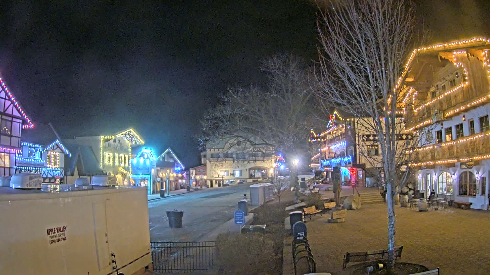 A town square at night, illuminated by colorful lights strung along buildings, under a clear, dark sky.