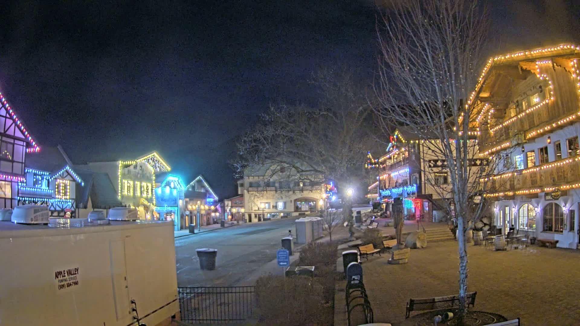 A nighttime scene of a town square, decorated with colorful Christmas lights on buildings and trees, with a few benches and a mostly empty street.
