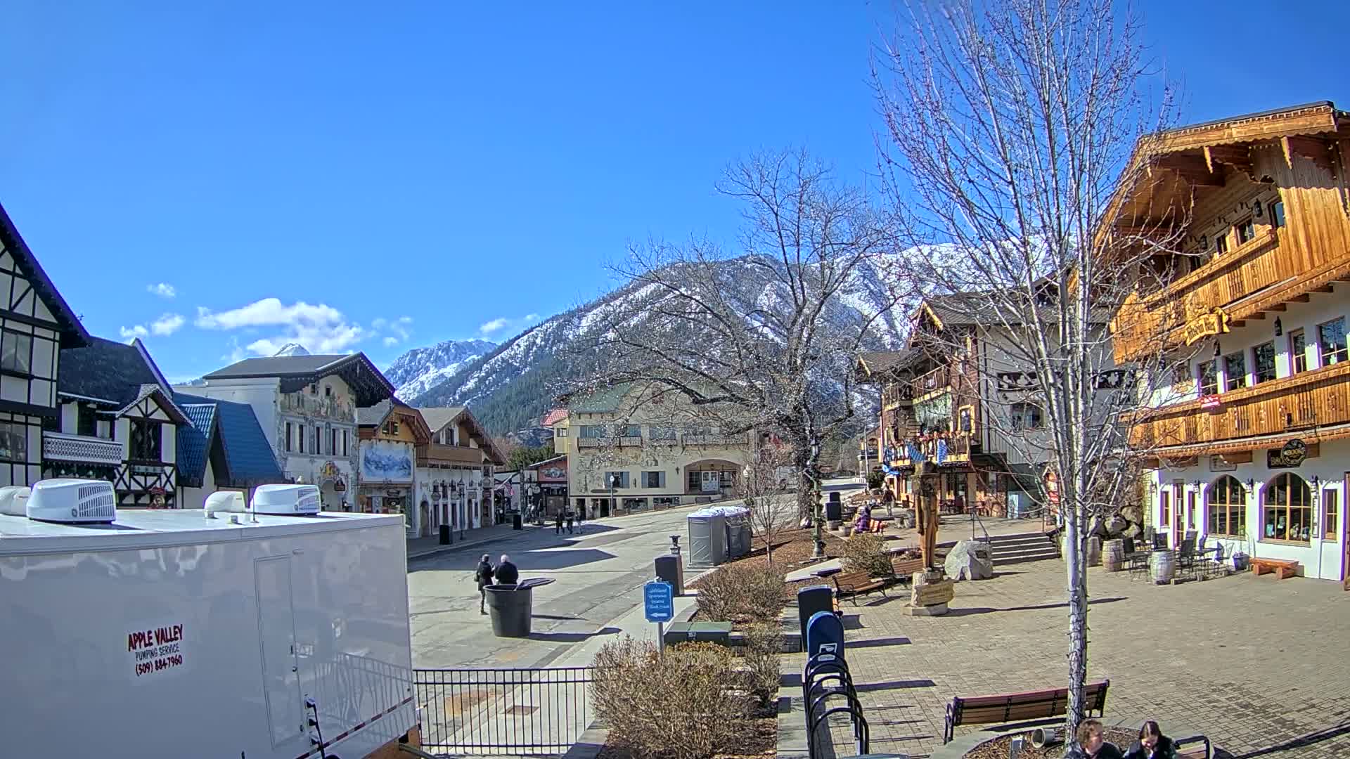 A sunny day reveals a Bavarian-style village square, with snow-capped mountains in the background, and a few pedestrians walking around.