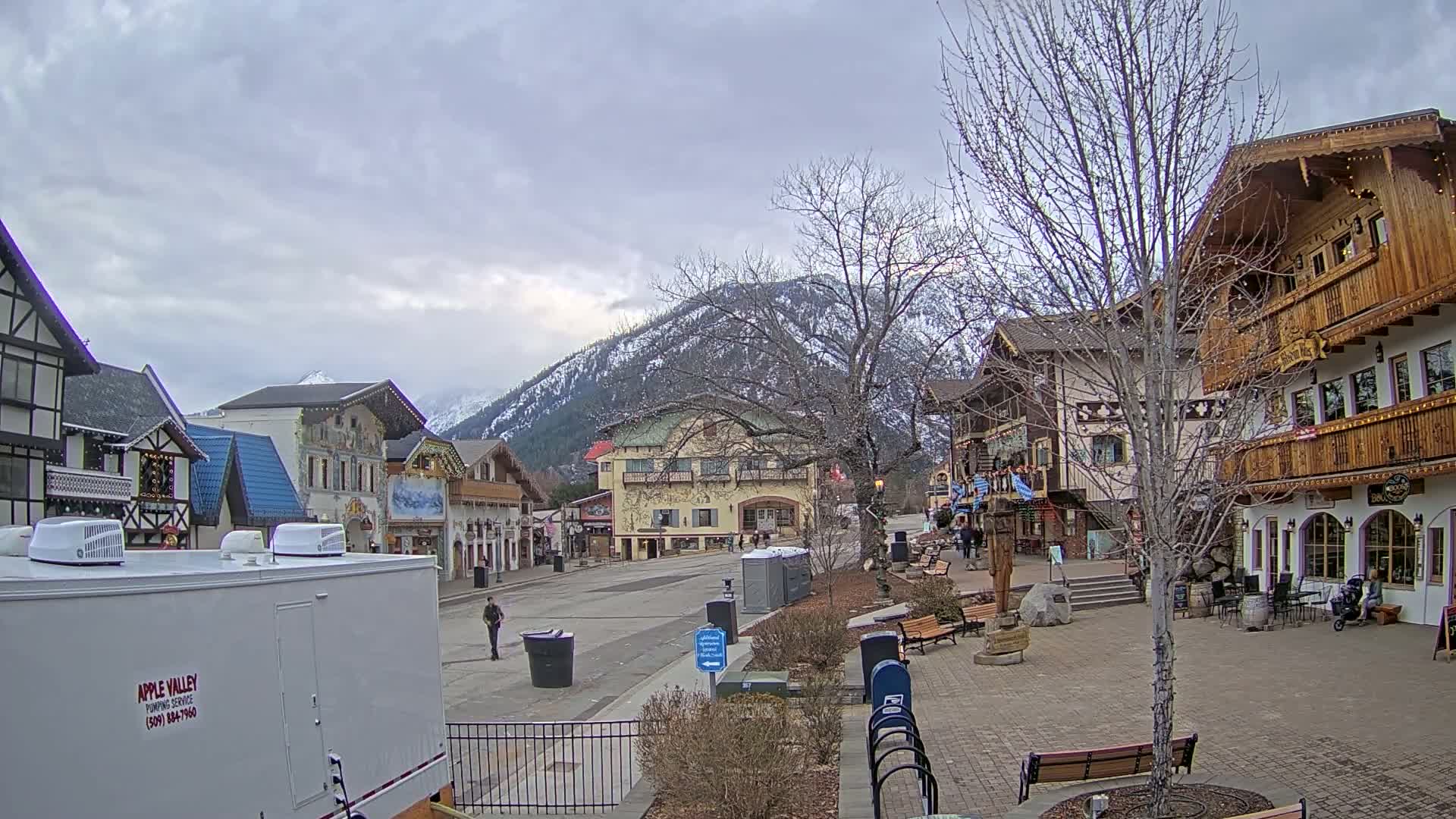 A mostly empty town square with European-style buildings, a few pedestrians, and snow-capped mountains in the background under a cloudy sky.