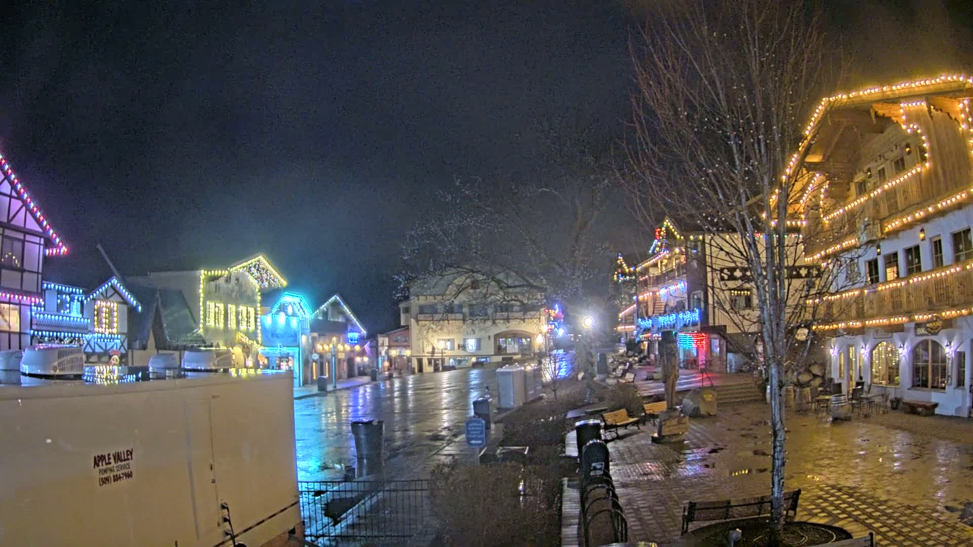 A town square at night, illuminated by colorful lights strung across buildings, is wet from recent rain.