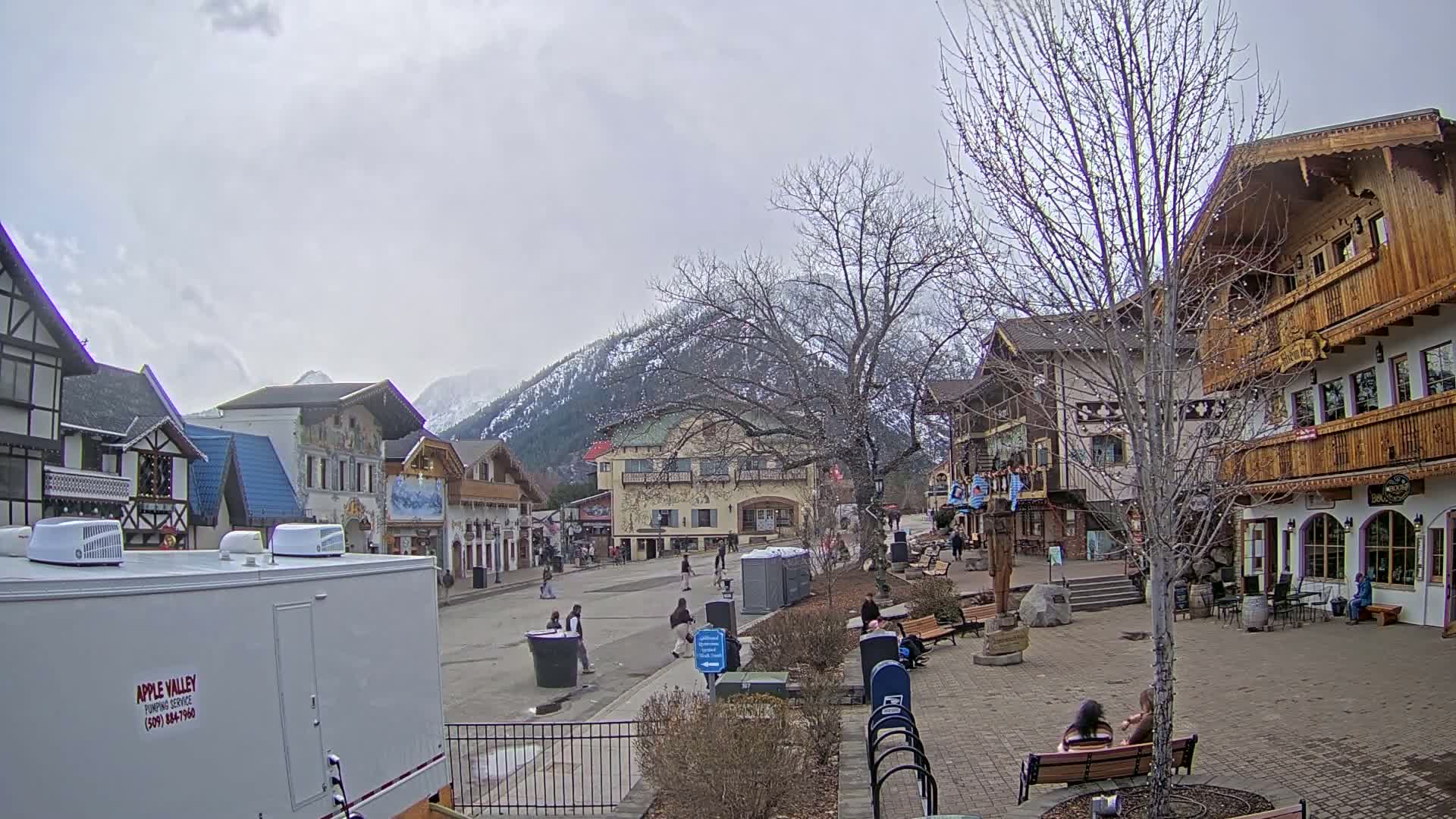 A mostly empty town square with European-style buildings and a mountain backdrop under an overcast sky.