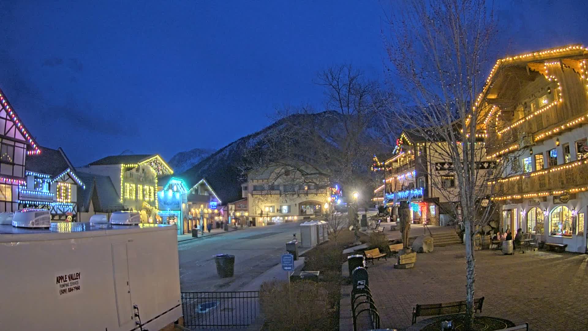 A town square at night, illuminated with colorful string lights on buildings, is nestled against a snow-dusted mountain under a dark blue, clear sky.