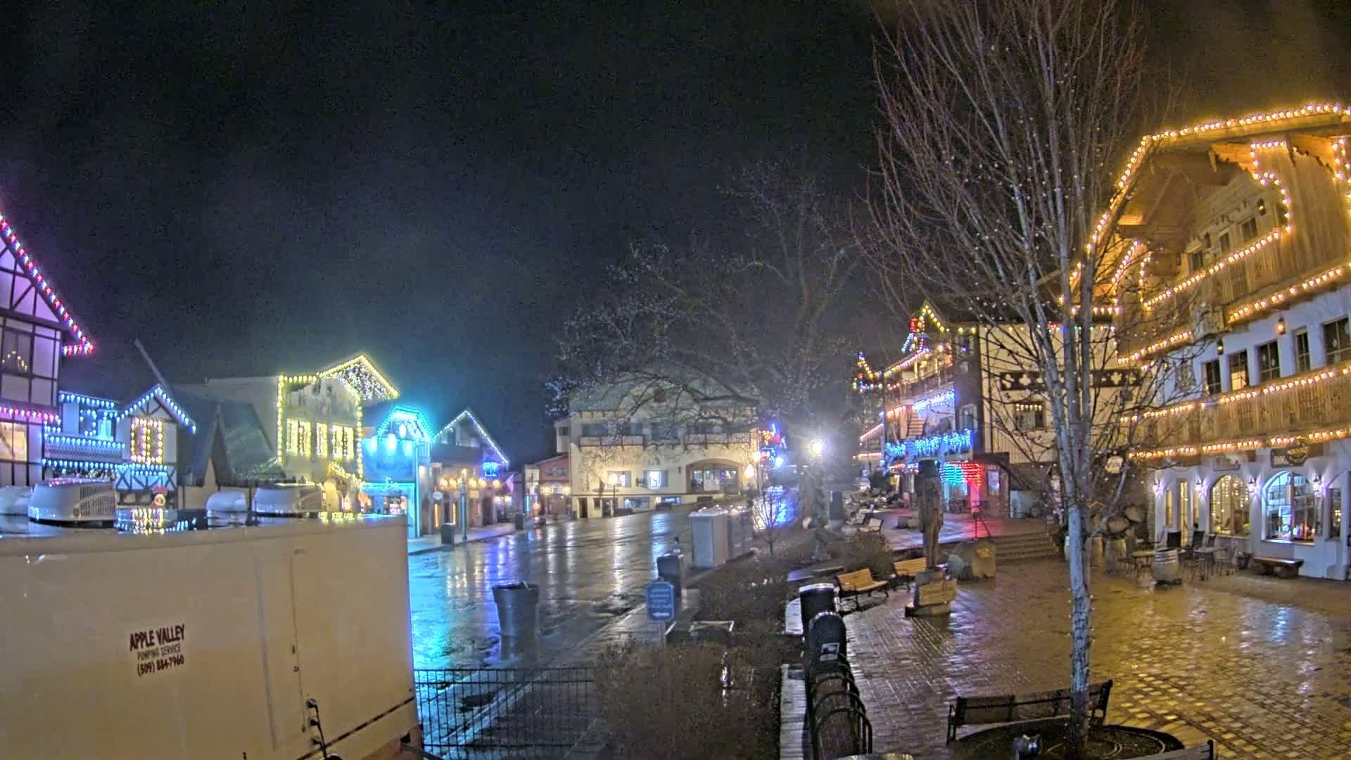 A town square at night, decorated with colorful lights on buildings, is wet from recent rain.