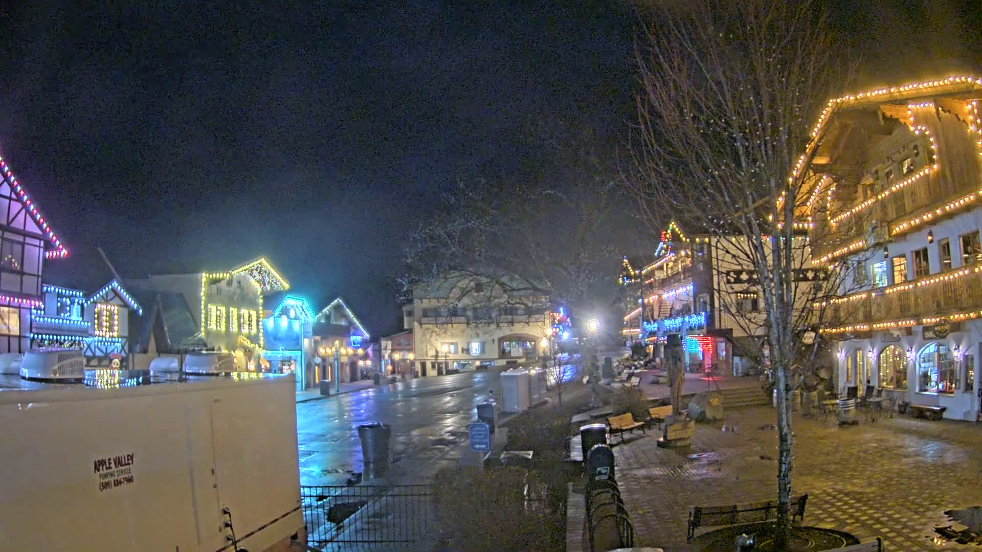 A town square at night, illuminated by colorful lights strung on buildings, is wet from recent rain or snow.