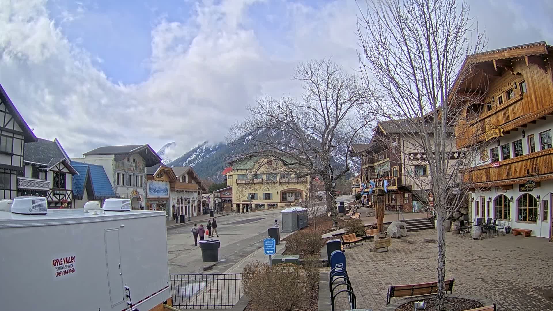 A partly cloudy day reveals a village of European-style buildings, a paved town square with benches, and snow-capped mountains in the distance.