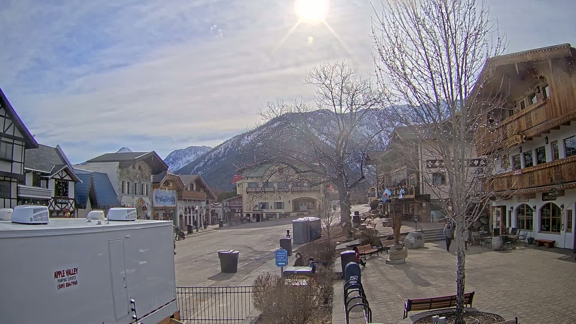 A mostly empty, quaint European-style town square sits before snow-capped mountains under a bright, sunny sky.
