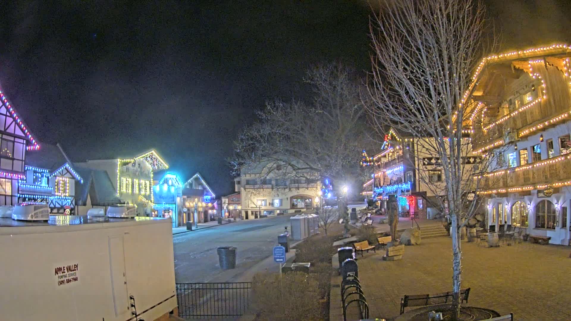 A nighttime scene of a village square with several buildings decorated with colorful lights, some snow on the ground, and bare trees.