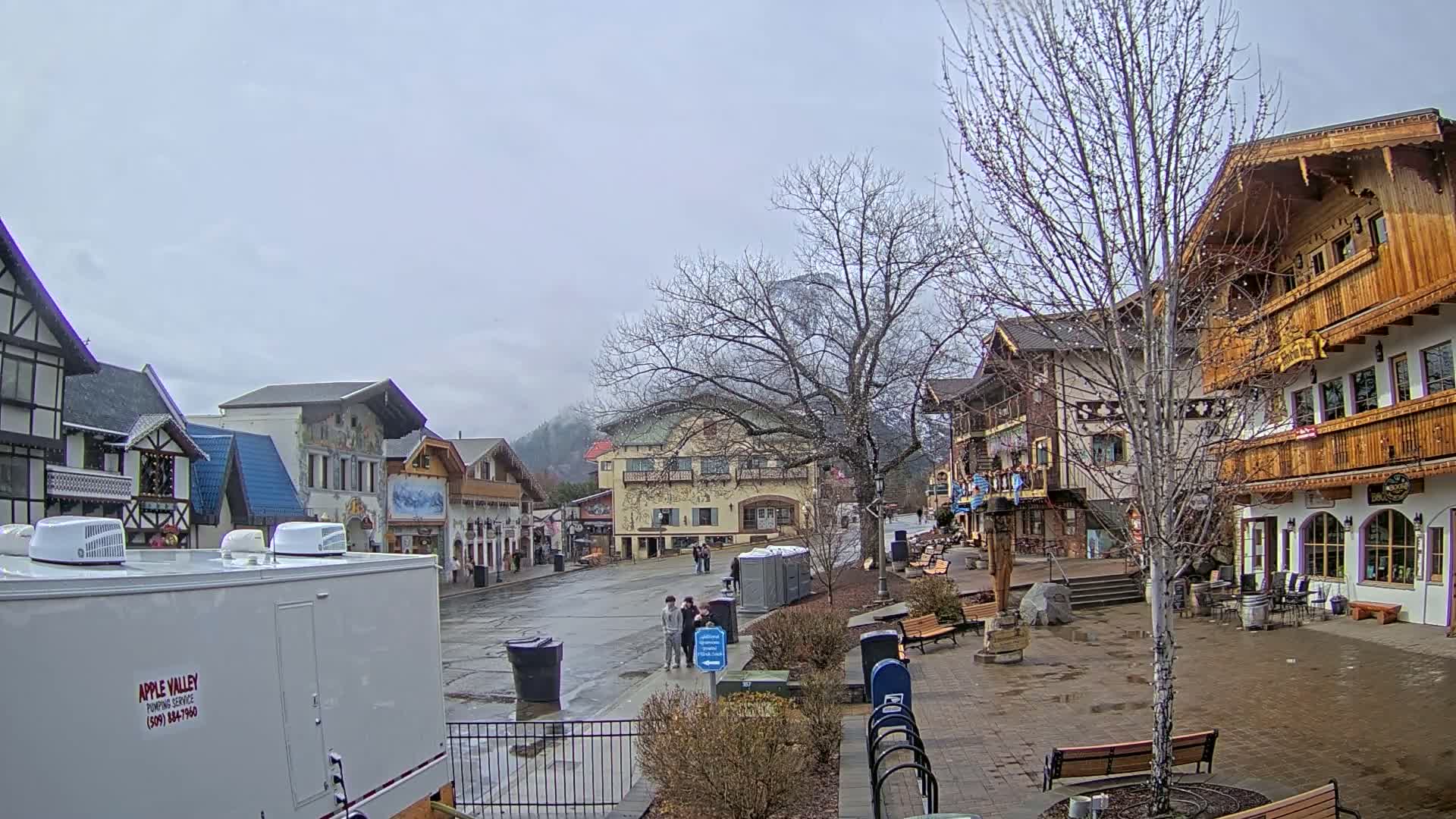 A wet, overcast day reveals a village square with several Bavarian-style buildings, a few pedestrians, and bare trees.