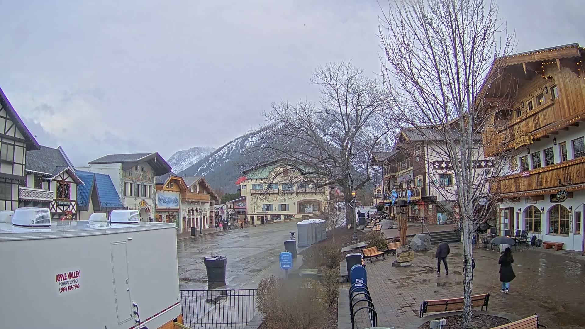 A wet town square with several buildings in an alpine style, set against a backdrop of snow-capped mountains under an overcast sky.