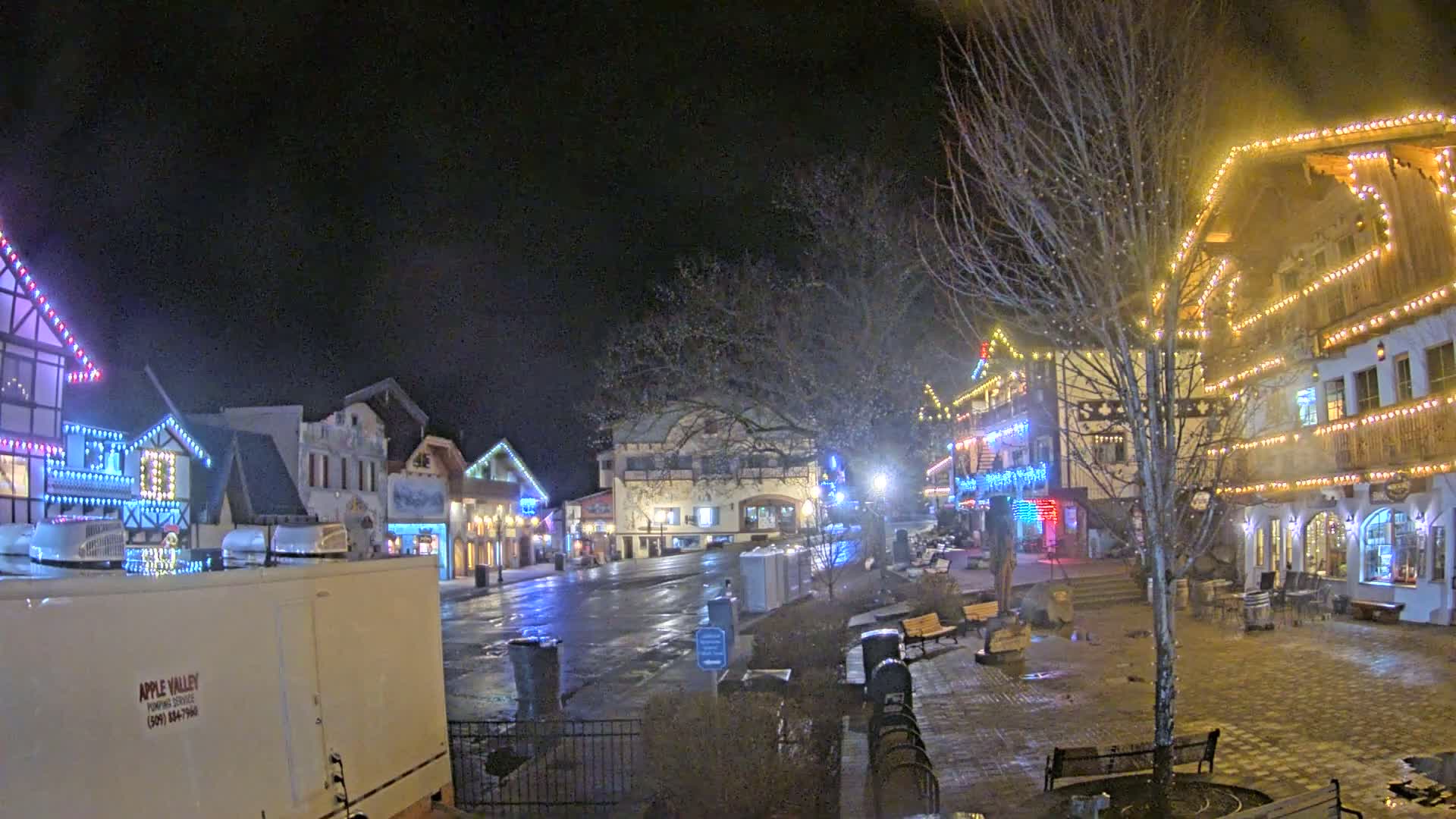 A town square at night, illuminated by colorful lights strung on buildings, shows a wet, possibly rainy or snowy, street.
