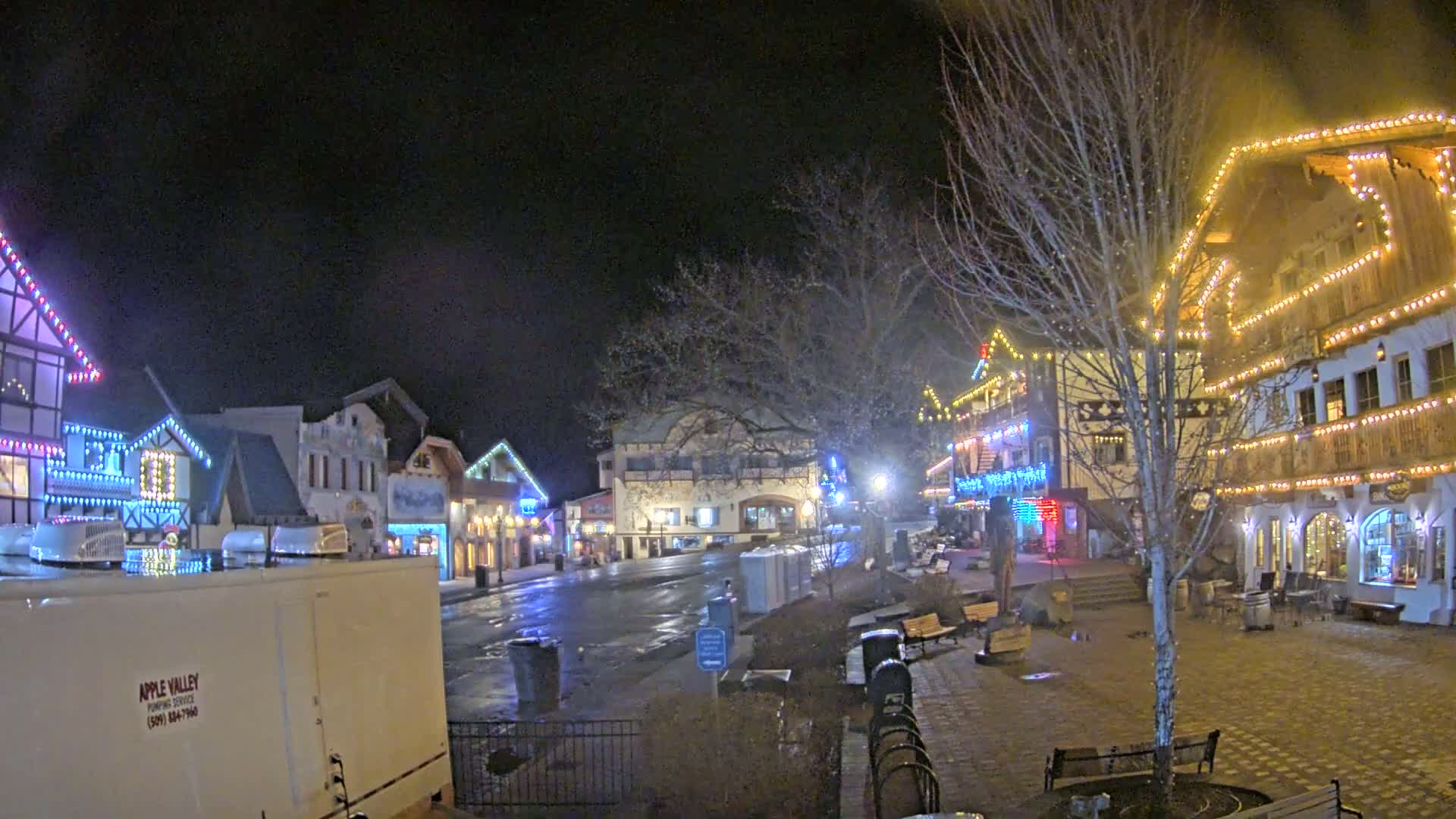 A town square at night, decorated with colorful Christmas lights, is wet from recent rain or snow.