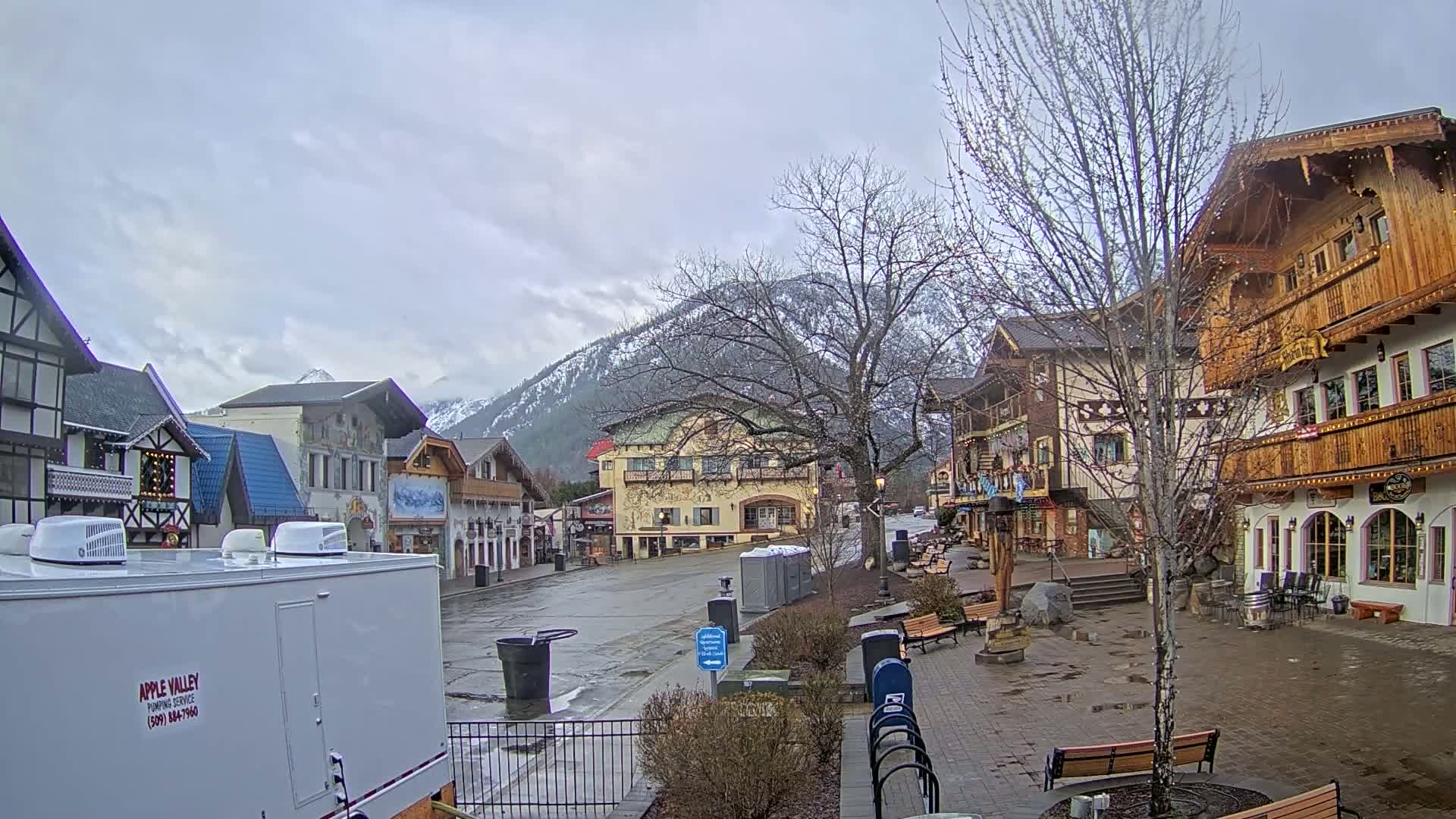 A mostly empty town square with Bavarian-style buildings, a snow-capped mountain in the background, and an overcast sky.