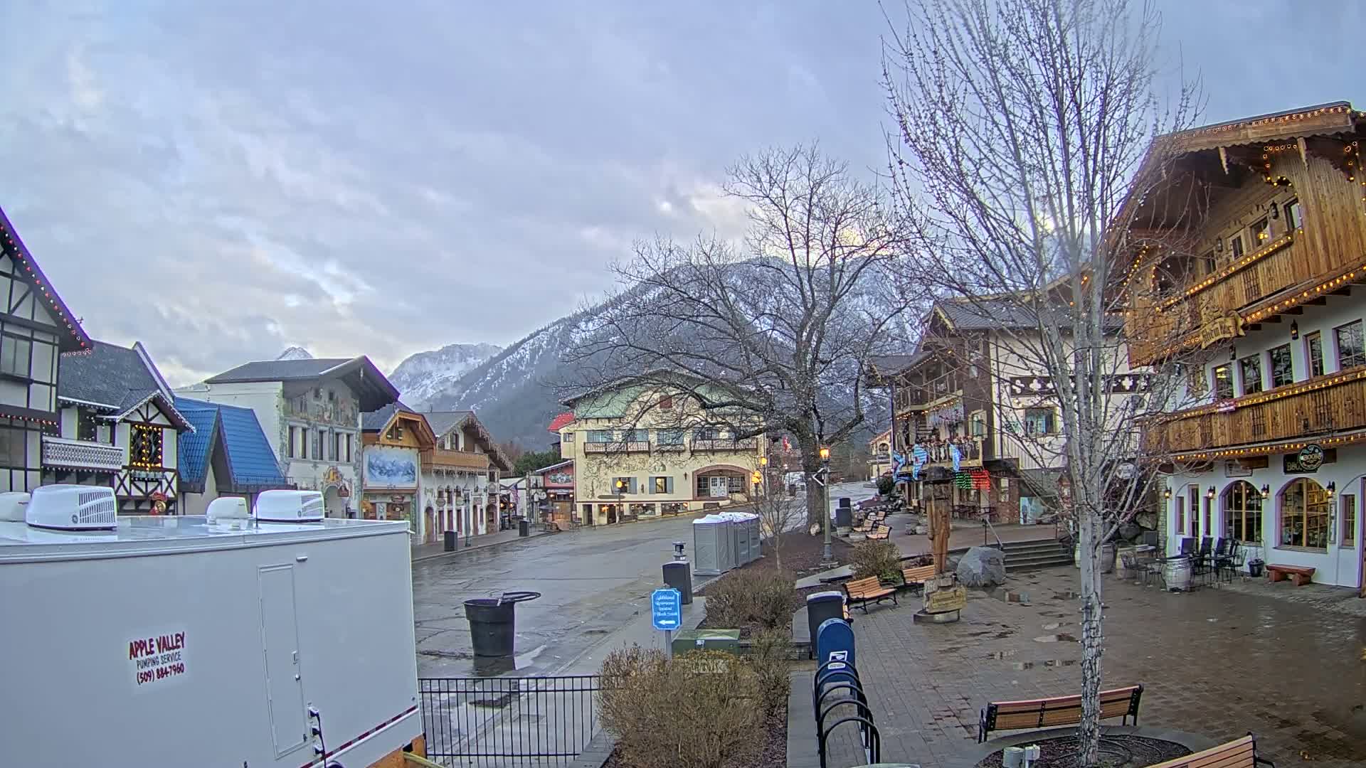 A town square with  European-style buildings, snow-capped mountains in the background, under a cloudy sky.