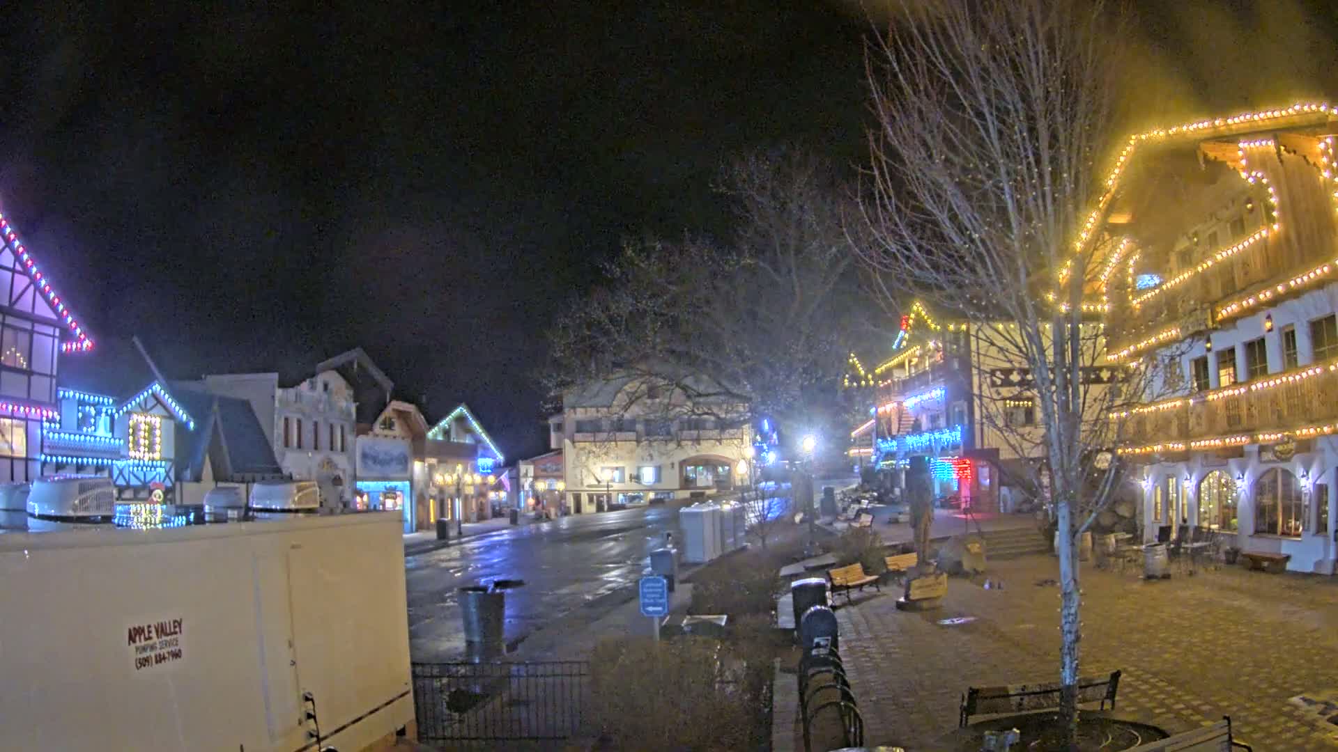 A town square at night, decorated with string lights on buildings and trees, shows a wet, paved area with benches and trash cans under a dark sky.