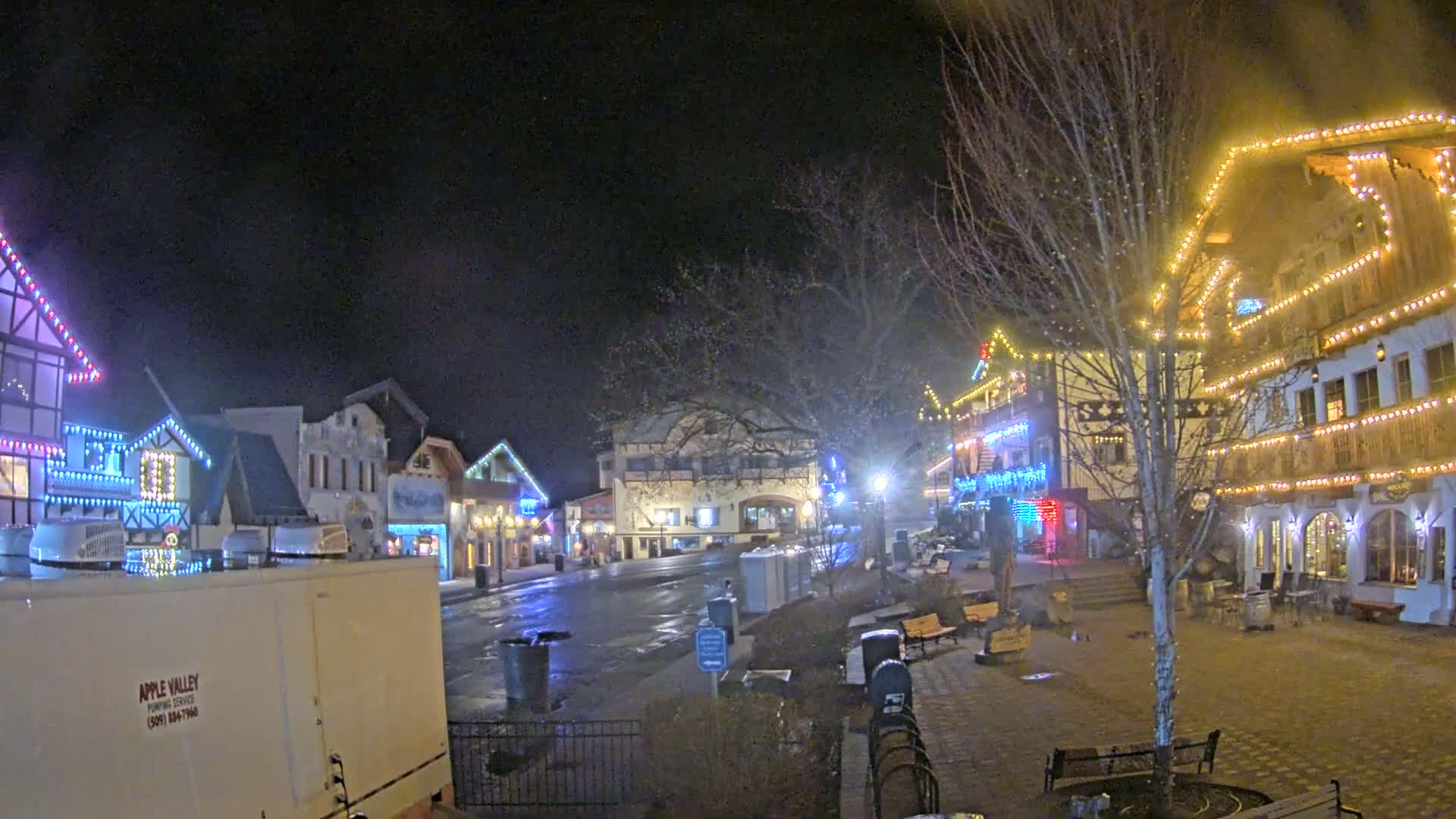 A nighttime view of a town square with several buildings decorated with Christmas lights, under a dark, possibly cloudy sky.