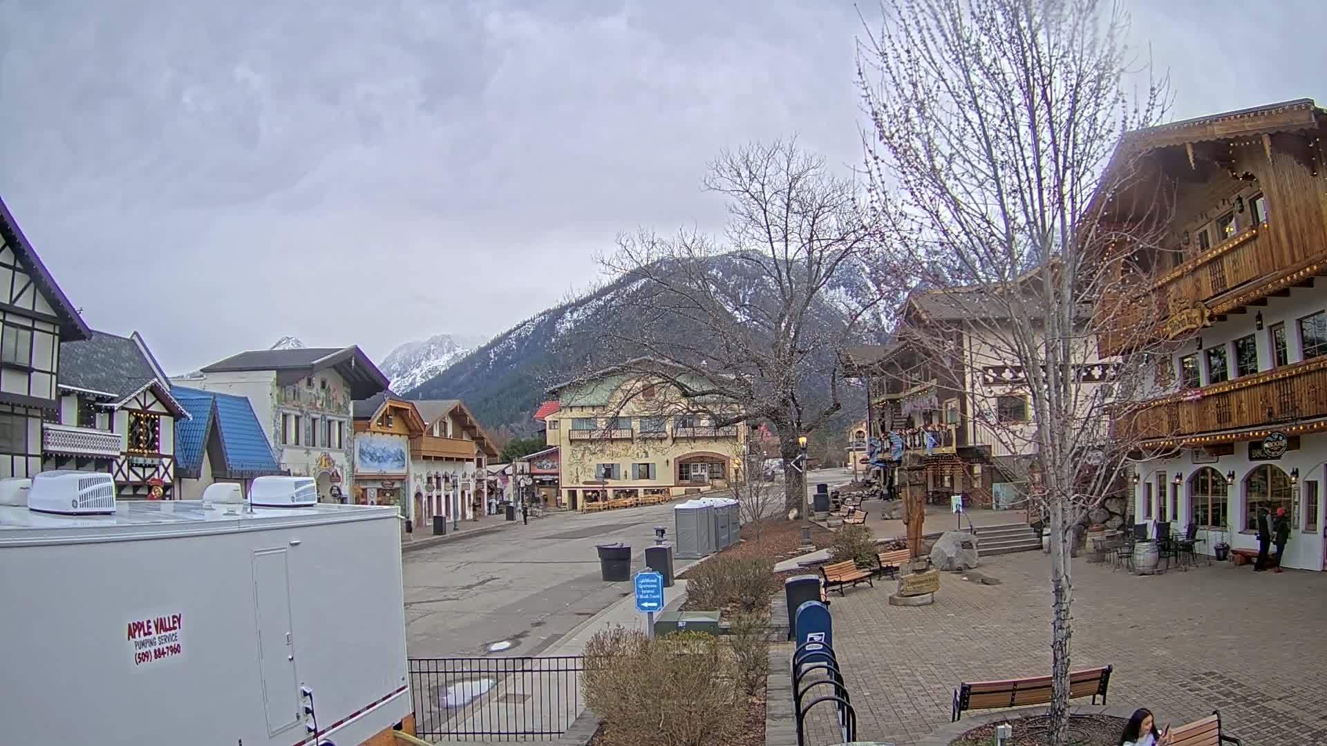 A mostly empty village square with several Bavarian-style buildings, a few benches, and a backdrop of snow-capped mountains under a cloudy sky.