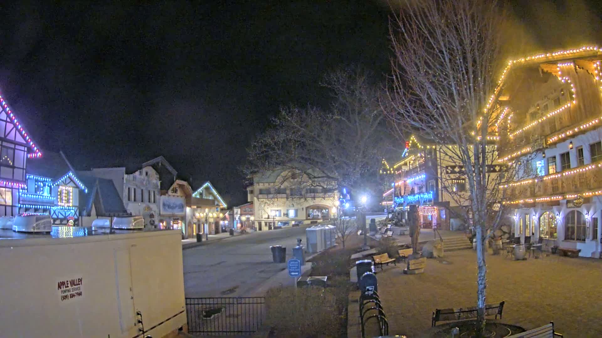 A nighttime scene of a village square with buildings decorated with Christmas lights, a few benches, and some small patches of snow on the ground.