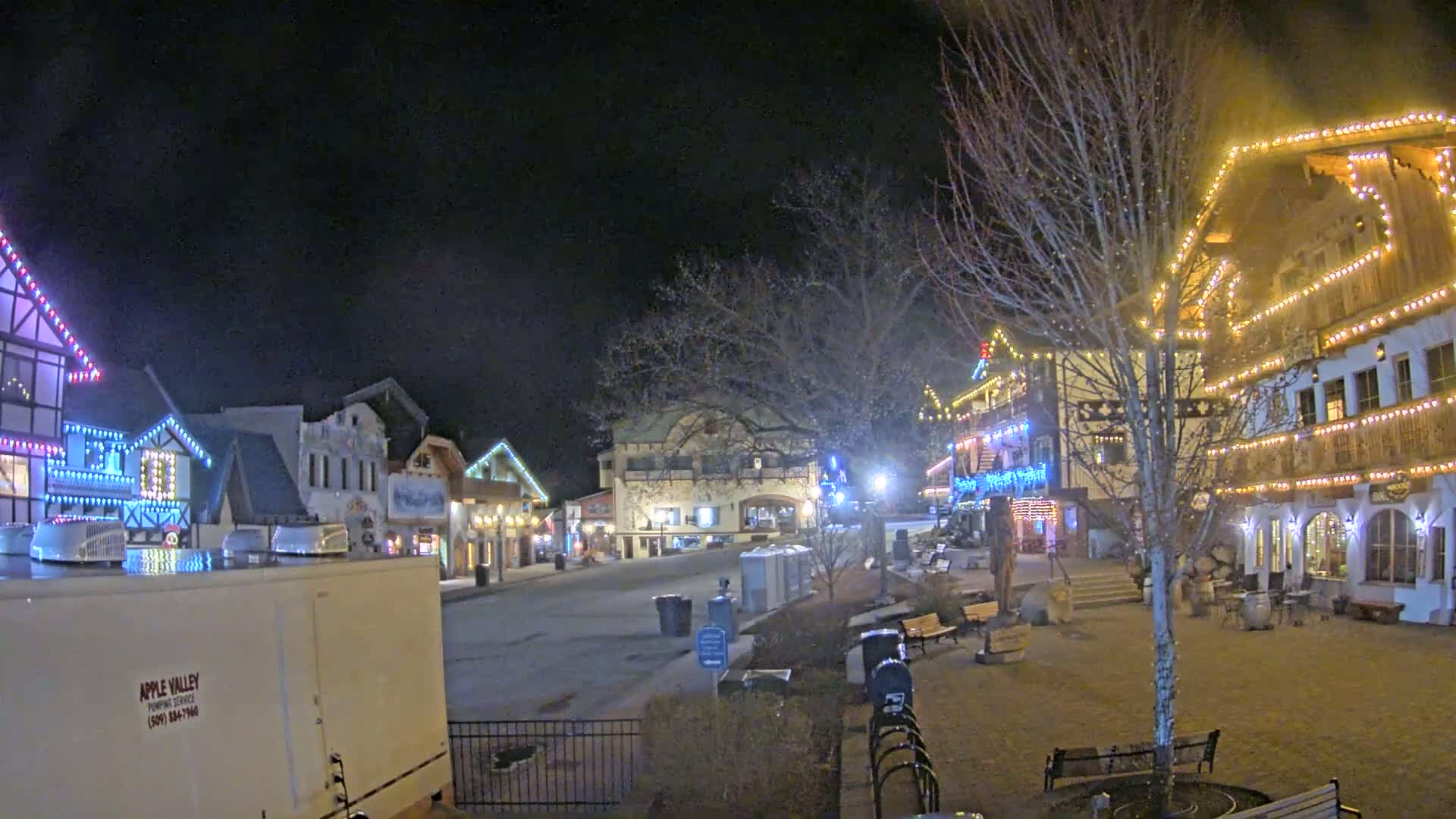 A nighttime scene of a village square with several buildings decorated with colorful lights, some snow on the ground, and bare trees.
