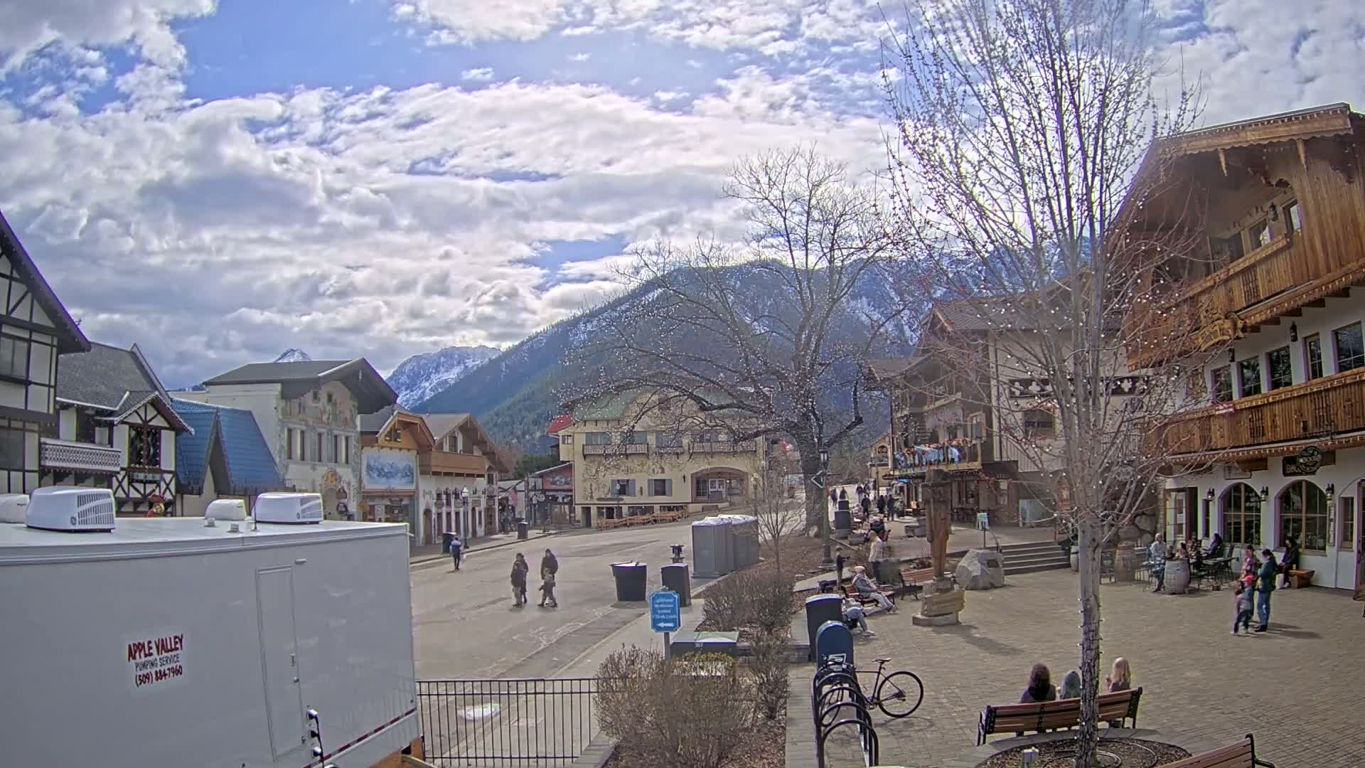 A partly cloudy day in a European-style village square nestled in a valley with mountains in the background, featuring several people walking and sitting around the plaza.