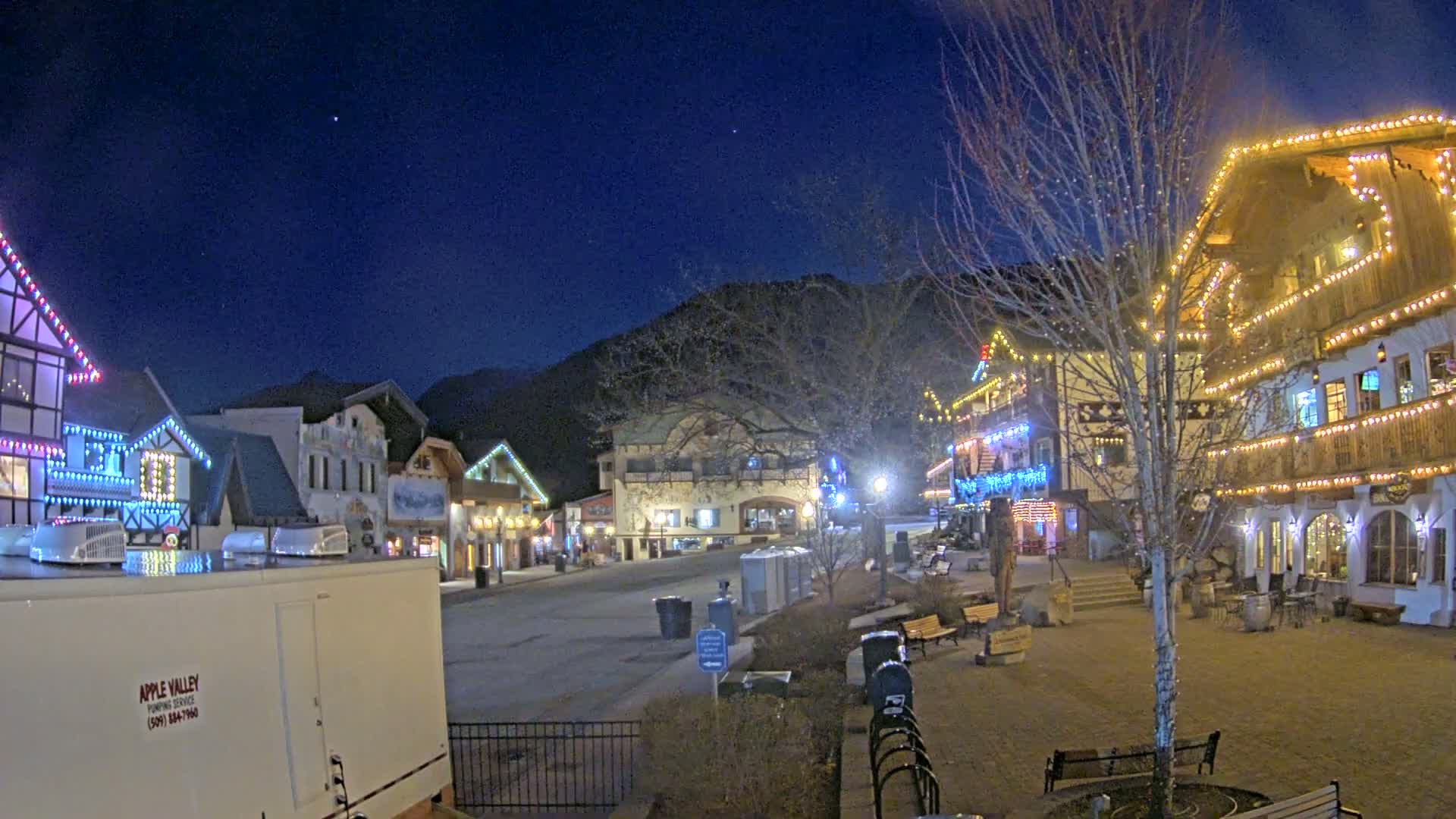 A nighttime view of a town square with buildings decorated with Christmas lights, under a clear, dark blue sky.