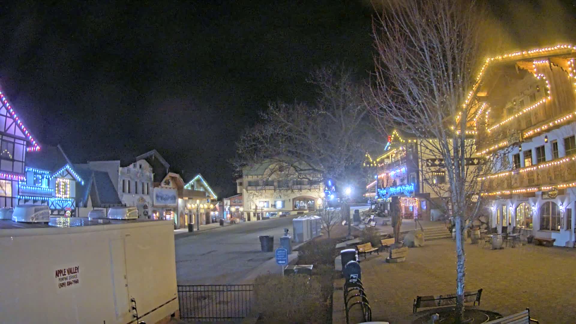 A nighttime view of a village square decorated with Christmas lights, featuring several buildings with festive lighting and some benches and landscaping.