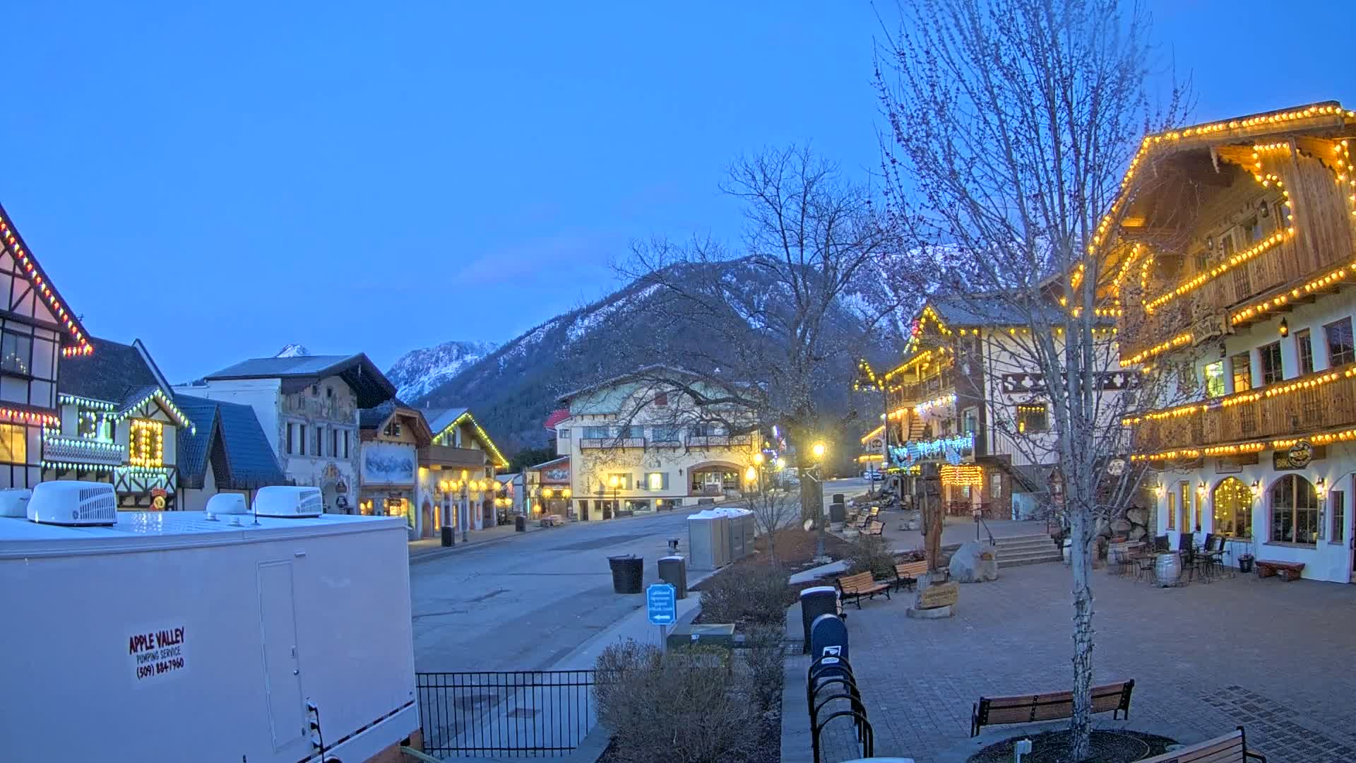 A quaint, festively lit town square nestled in a mountain valley under a twilight sky.