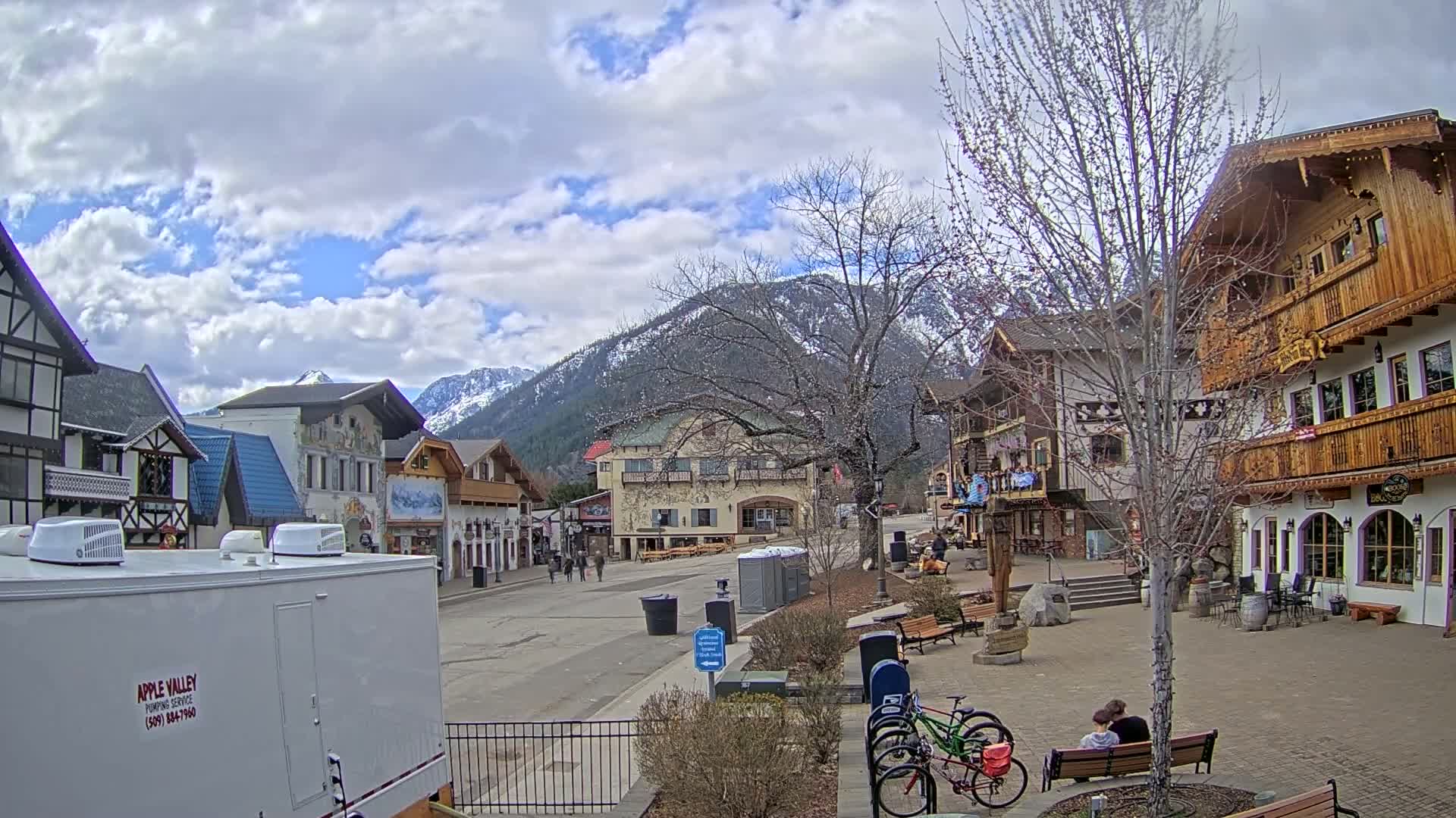 A partly cloudy day reveals a village square with Bavarian-style buildings, a mountain backdrop, and a few people and bicycles.