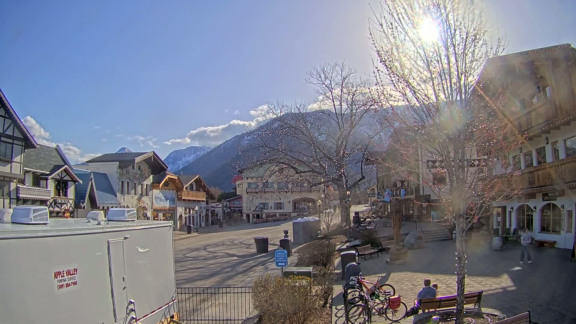 A sunny day in a village square with European-style buildings, a snow-capped mountain range in the background, and a few people sitting on a bench.