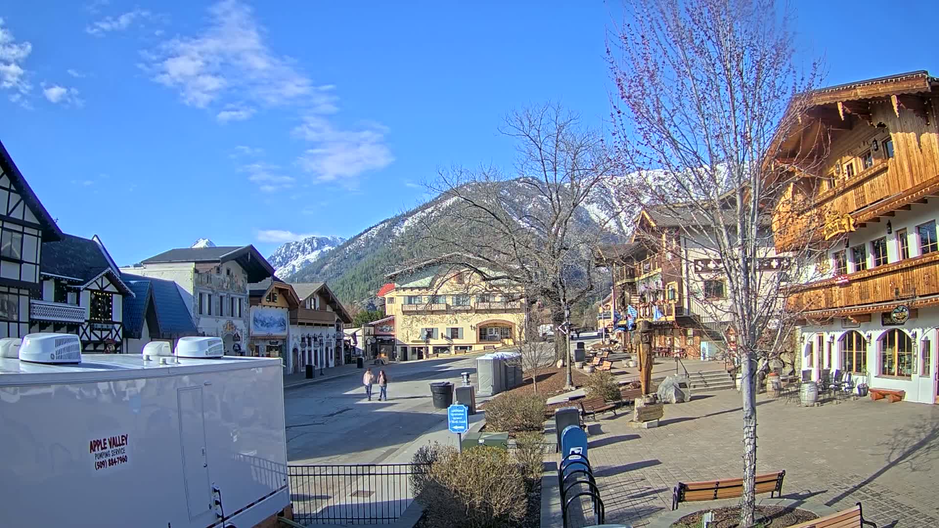 A partly sunny day reveals a quaint village square nestled in a valley, surrounded by buildings of European-inspired architecture and snow-capped mountains in the distance.