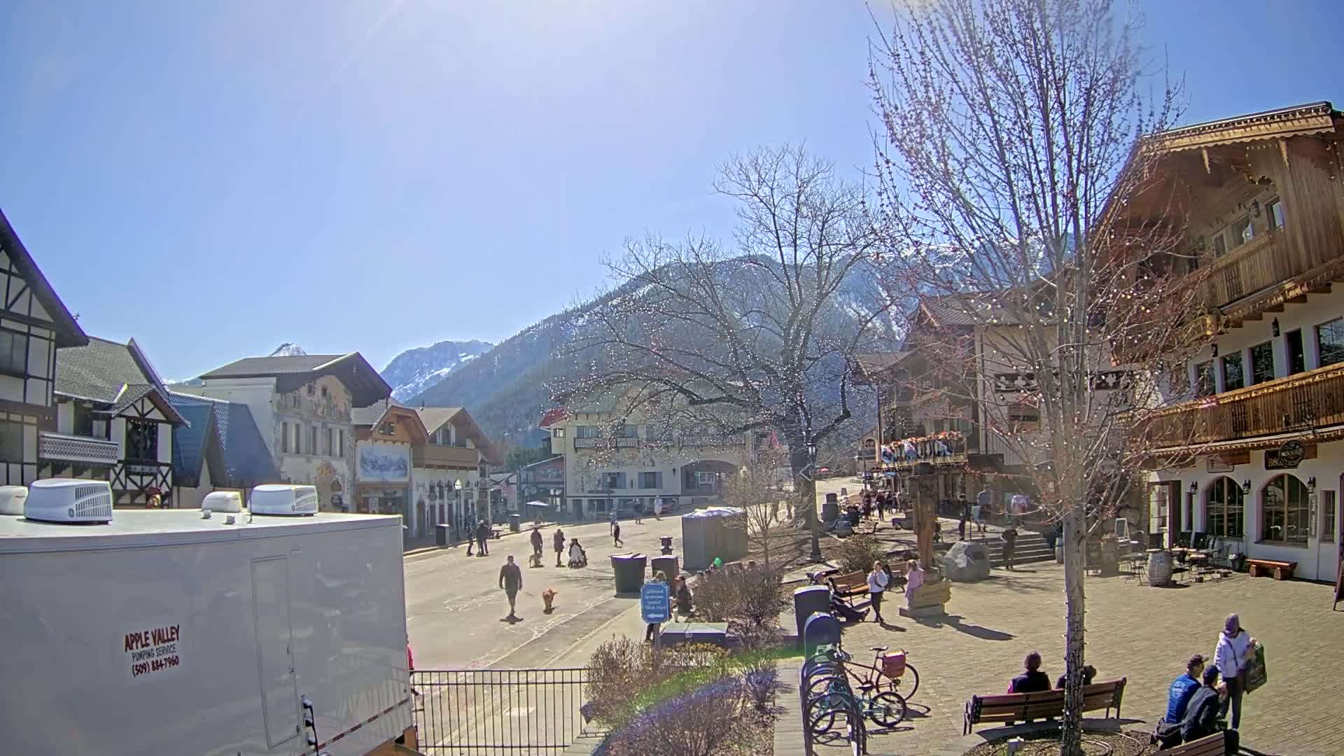 A sunny day in a village square with European-style buildings, people walking and sitting, and mountains in the background.