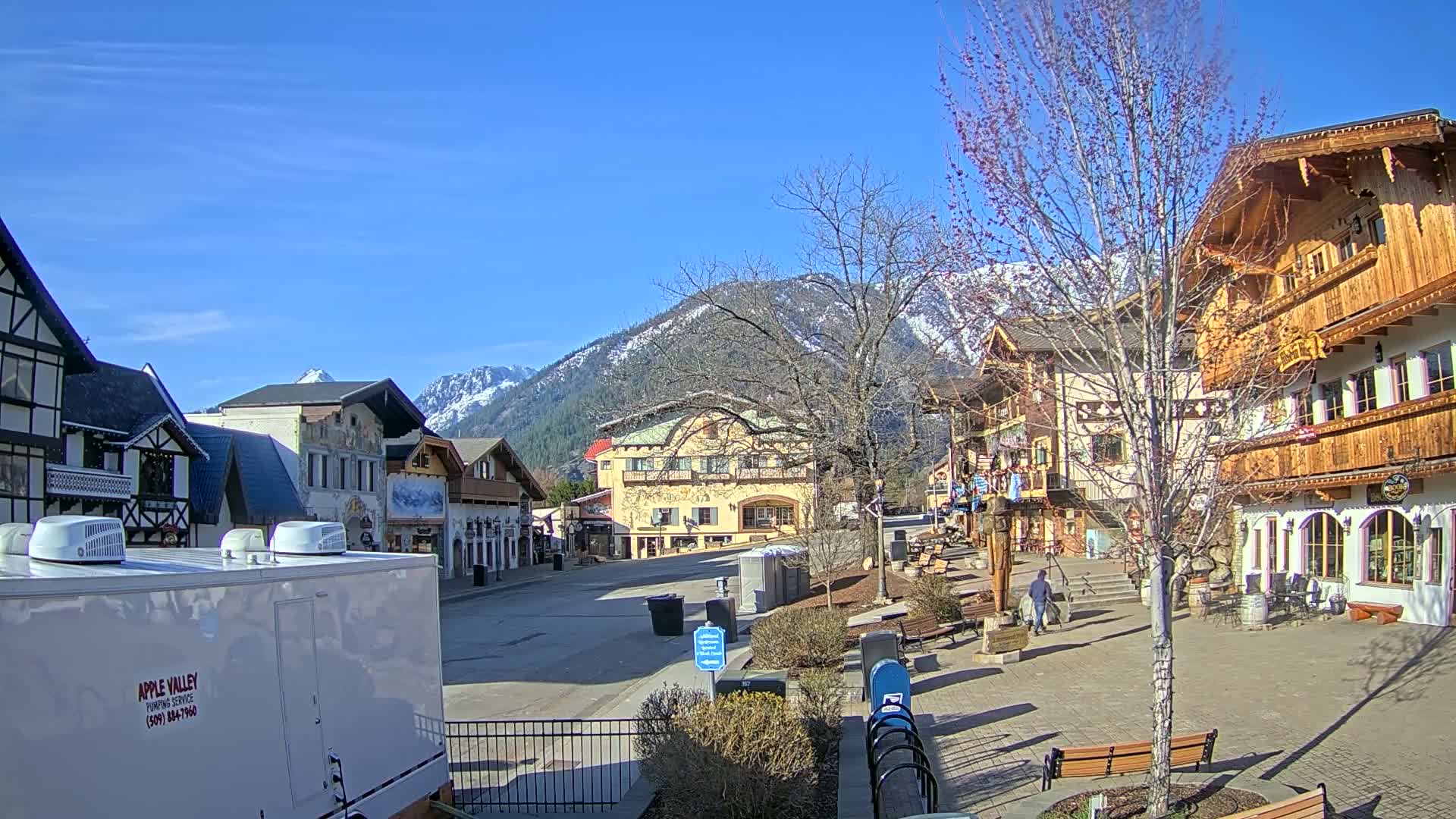 A sunny day reveals a quaint village square nestled against snow-capped mountains, with several buildings in European architectural styles.